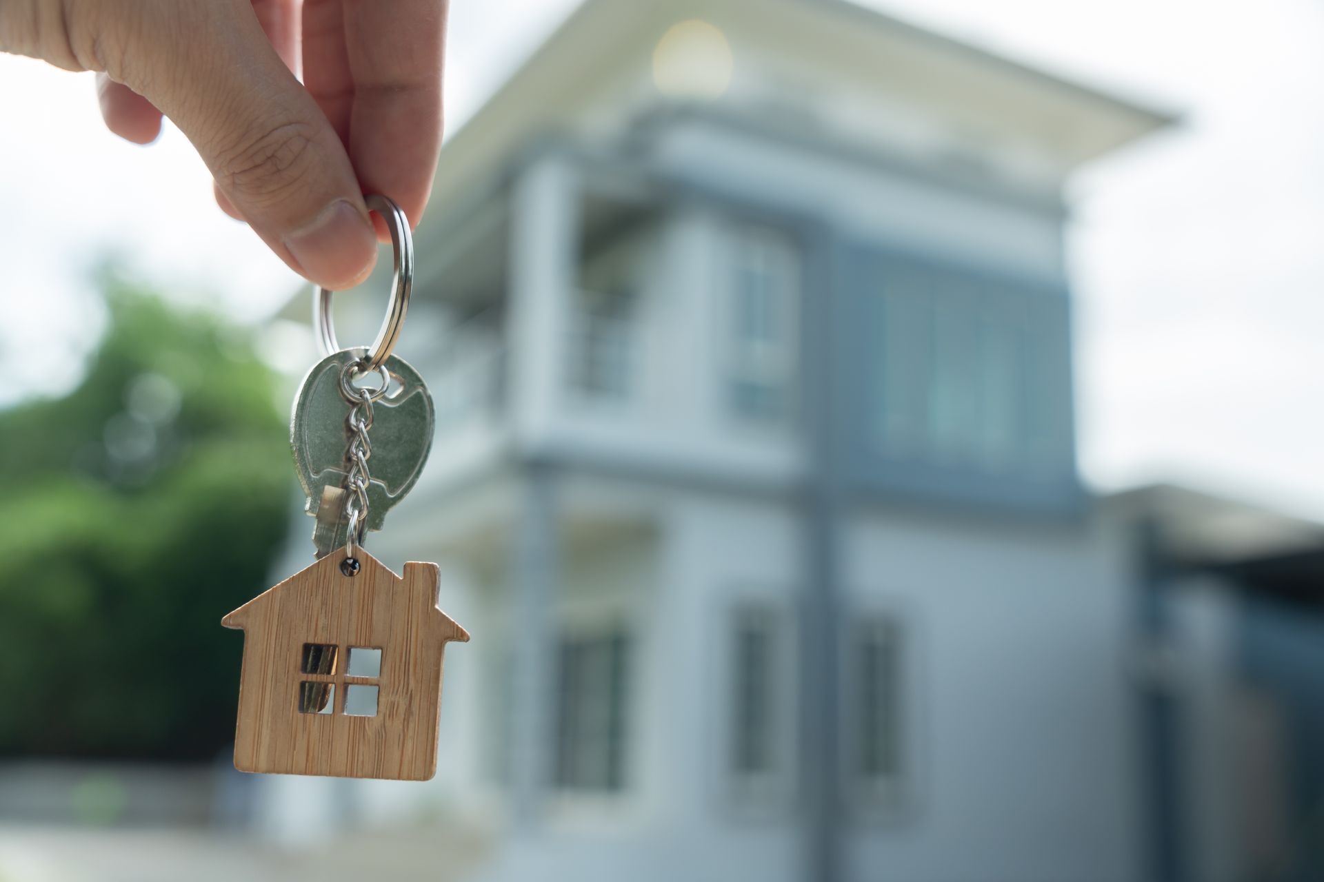 Hand holding keys with house-shaped charm in front of a blurred house.