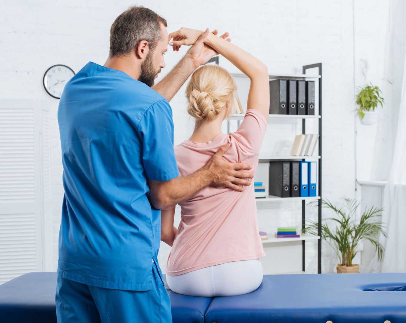 A man is helping a woman stretch her arms while sitting on a table