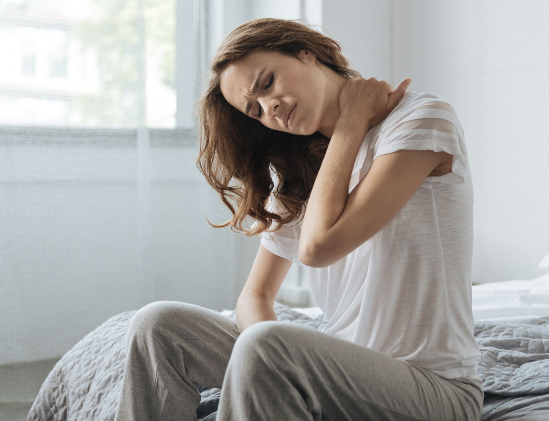 A woman is sitting on a bed holding her neck in pain
