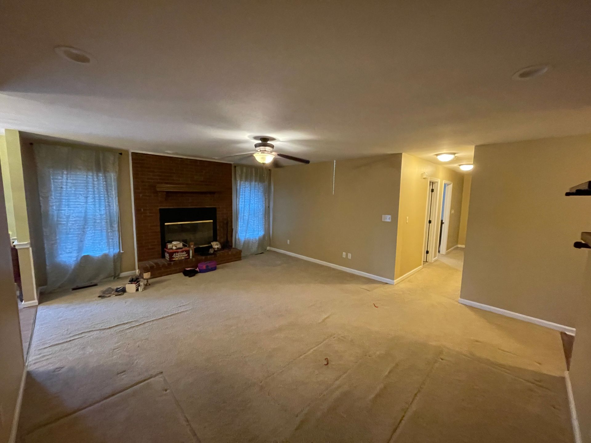 Spacious living room with a fireplace, beige walls, and light-colored carpet. Curtains flank the windows.