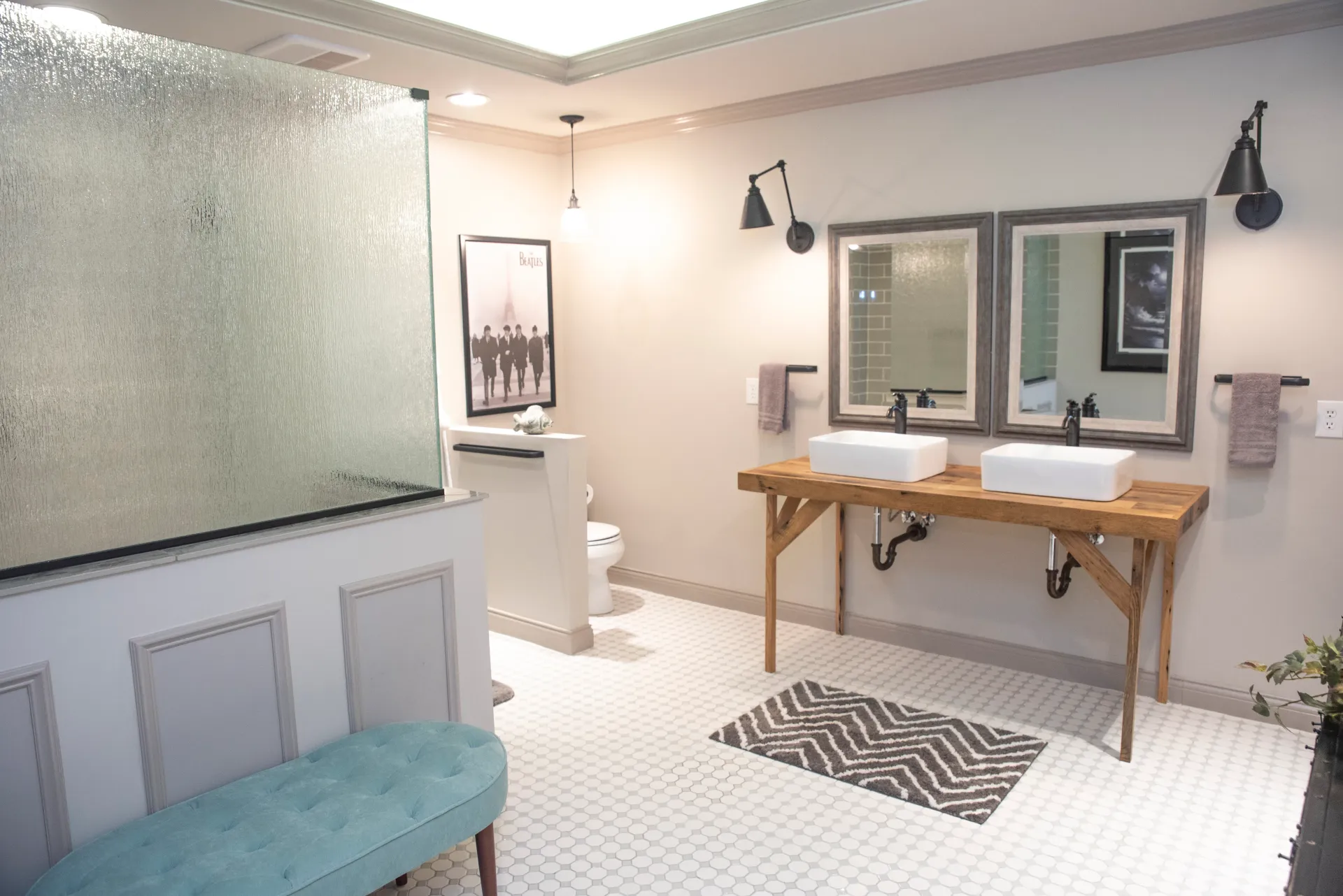 Bathroom with white tiled floor, double sinks on wooden table, glass shower, and turquoise bench.