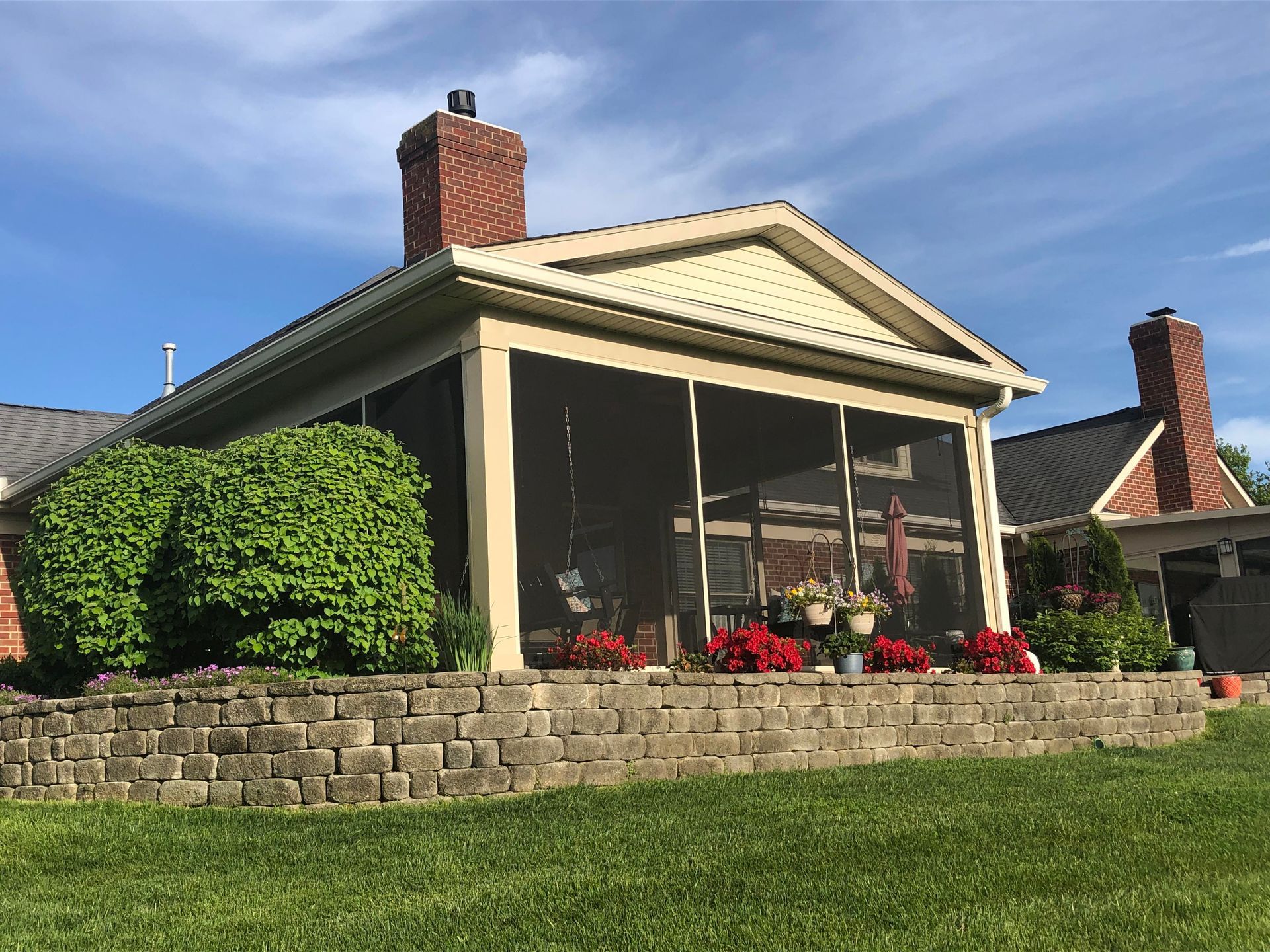 Screened porch with brick chimney and stonewall, red flowers, surrounded by grass.