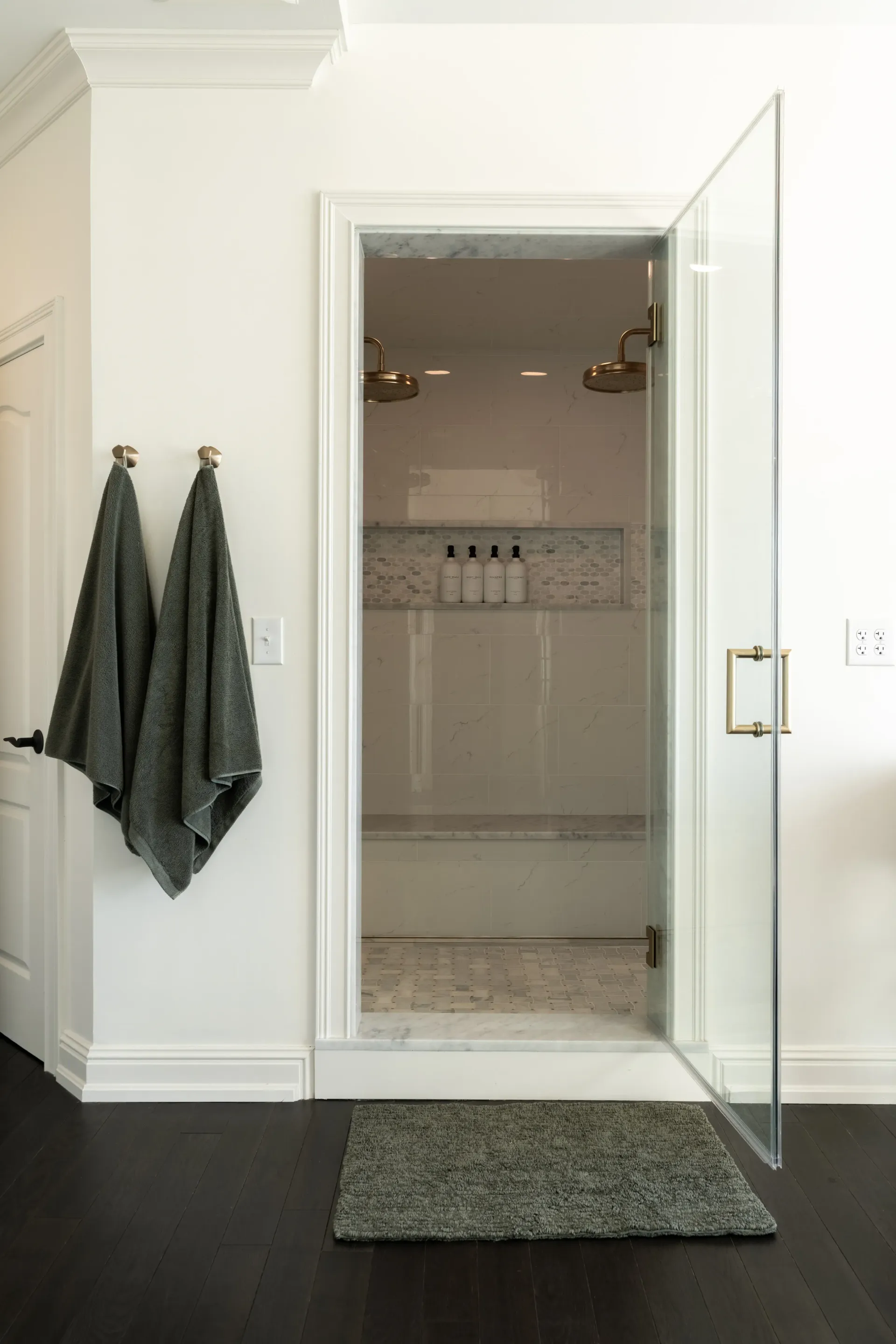 Bathroom with shower; glass door open, two showerheads visible, dark floor, towels on hooks.