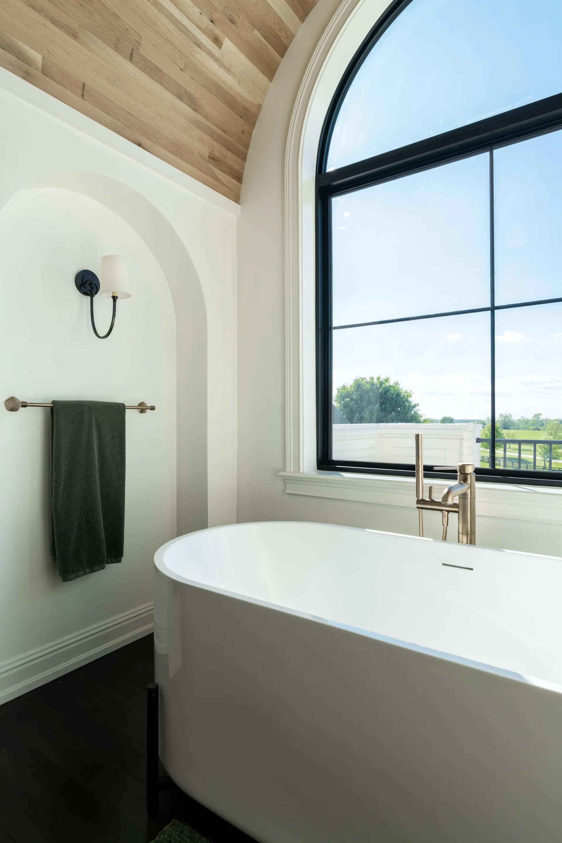 Bathroom with a white oval tub, large arched window, and wood ceiling. A towel hangs near a wall sconce.