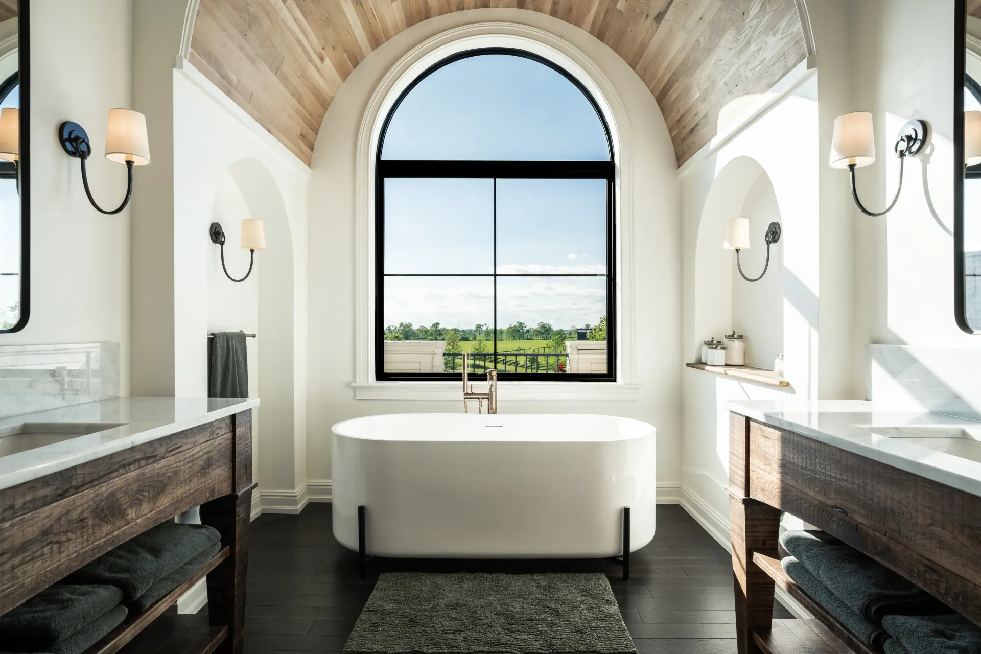 Bathroom with arched window, freestanding tub, dark wood vanities, sconce lighting, and wood ceiling.
