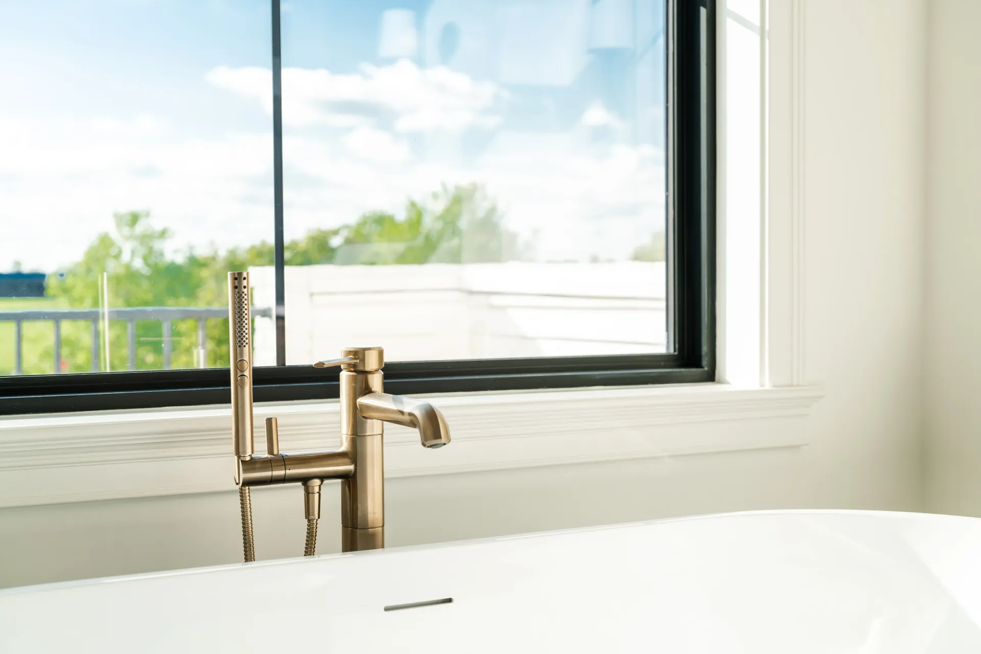 Close-up of a bathtub with gold faucet in front of a window. Exterior view of trees and sky visible.