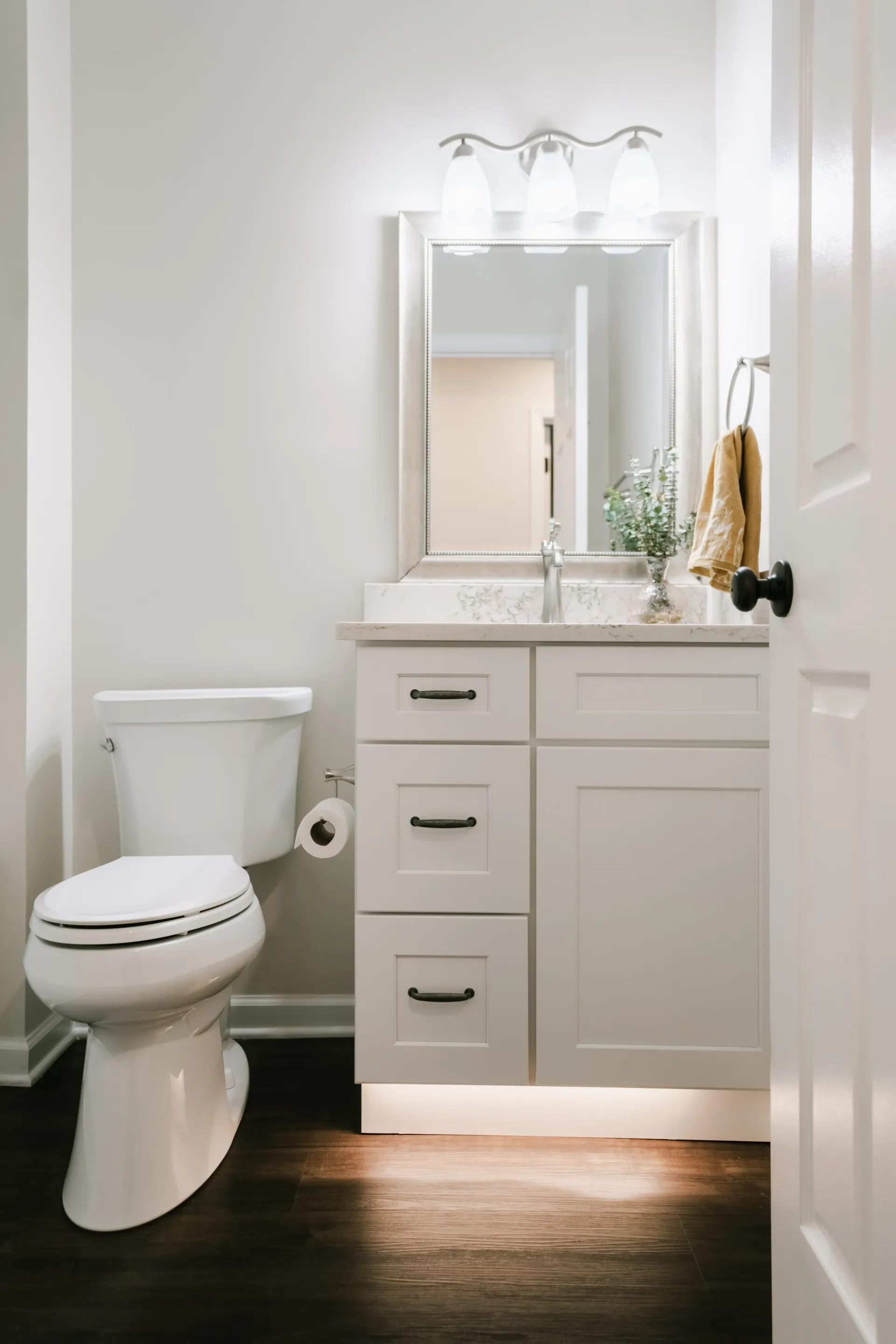 Bathroom with white vanity, toilet, and mirror, dark wood floor, and white walls.
