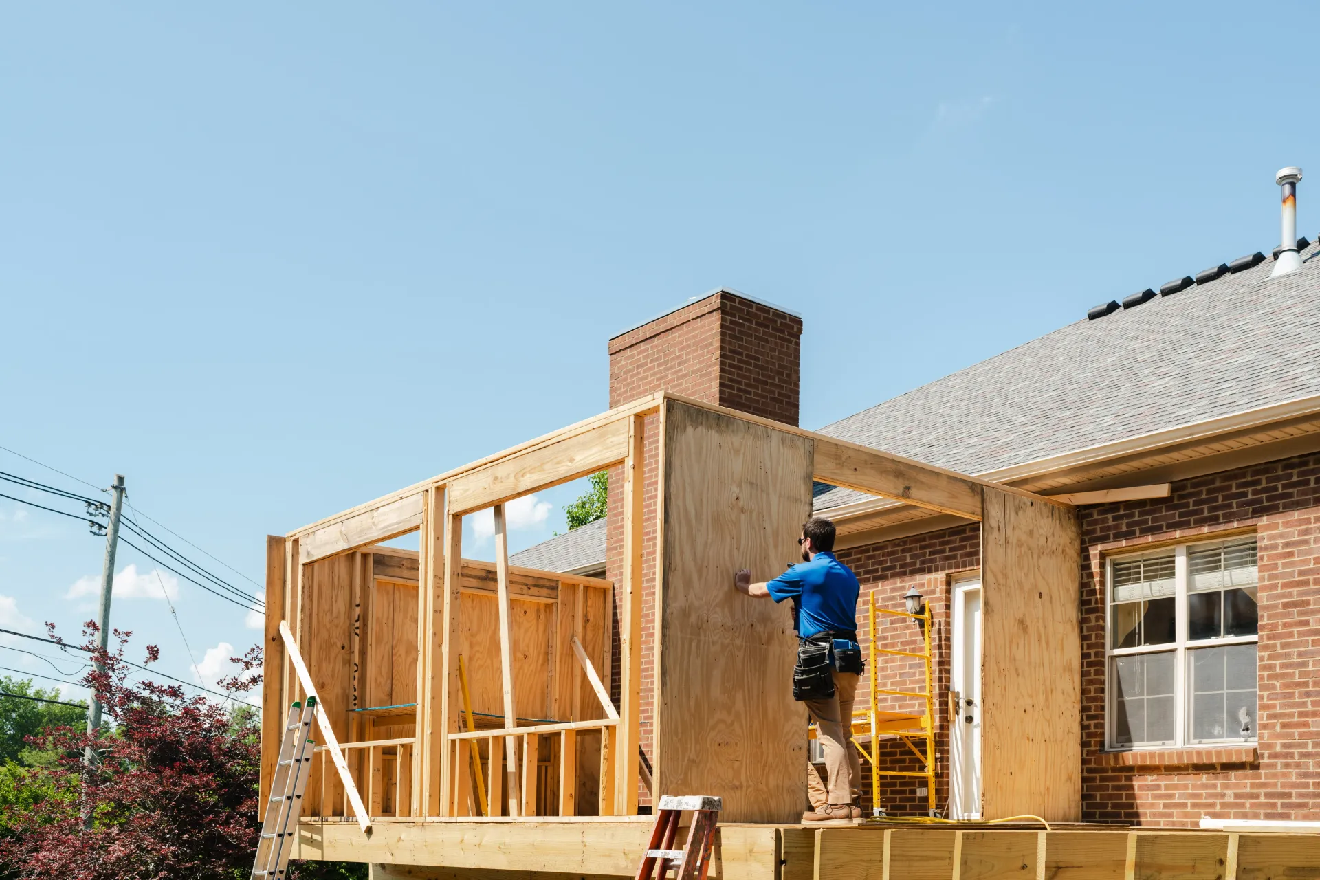 Construction worker framing a wooden structure attached to a brick house. Sunny day.