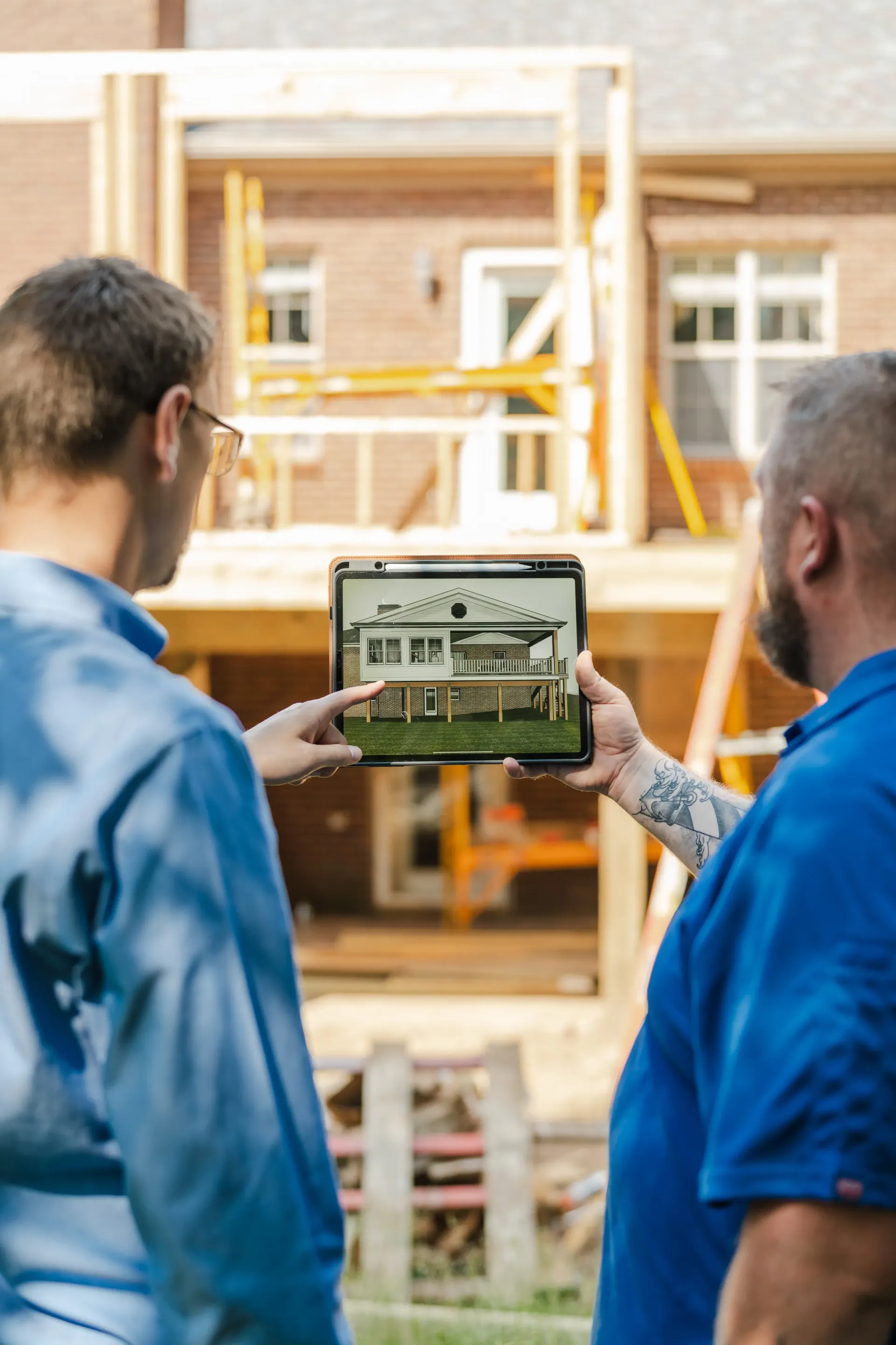 Two men in blue shirts examine a tablet displaying a house design, construction site in the background.