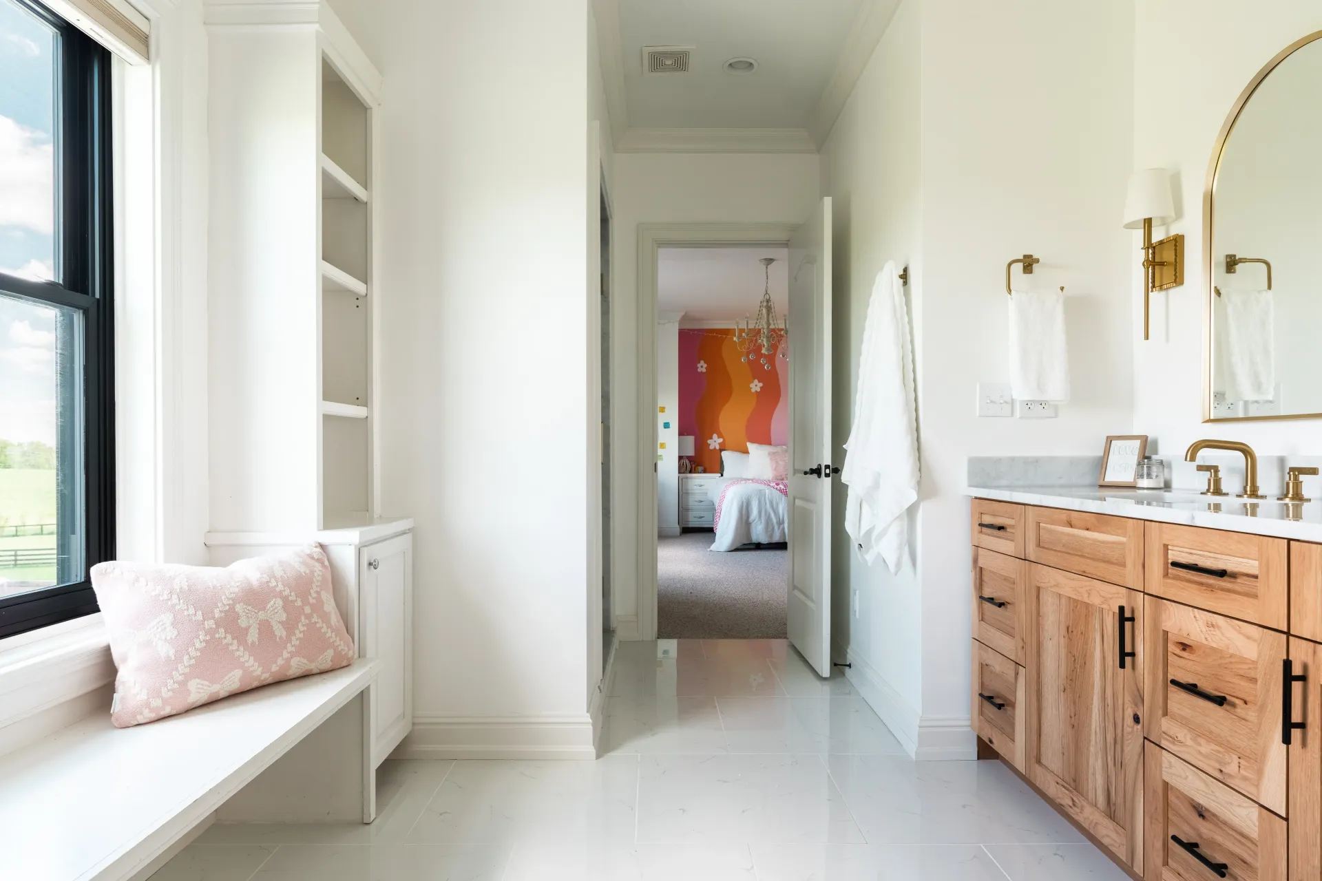 Bathroom with window seat, vanity, and hallway to bedroom. White walls, wood cabinets, and gold fixtures.