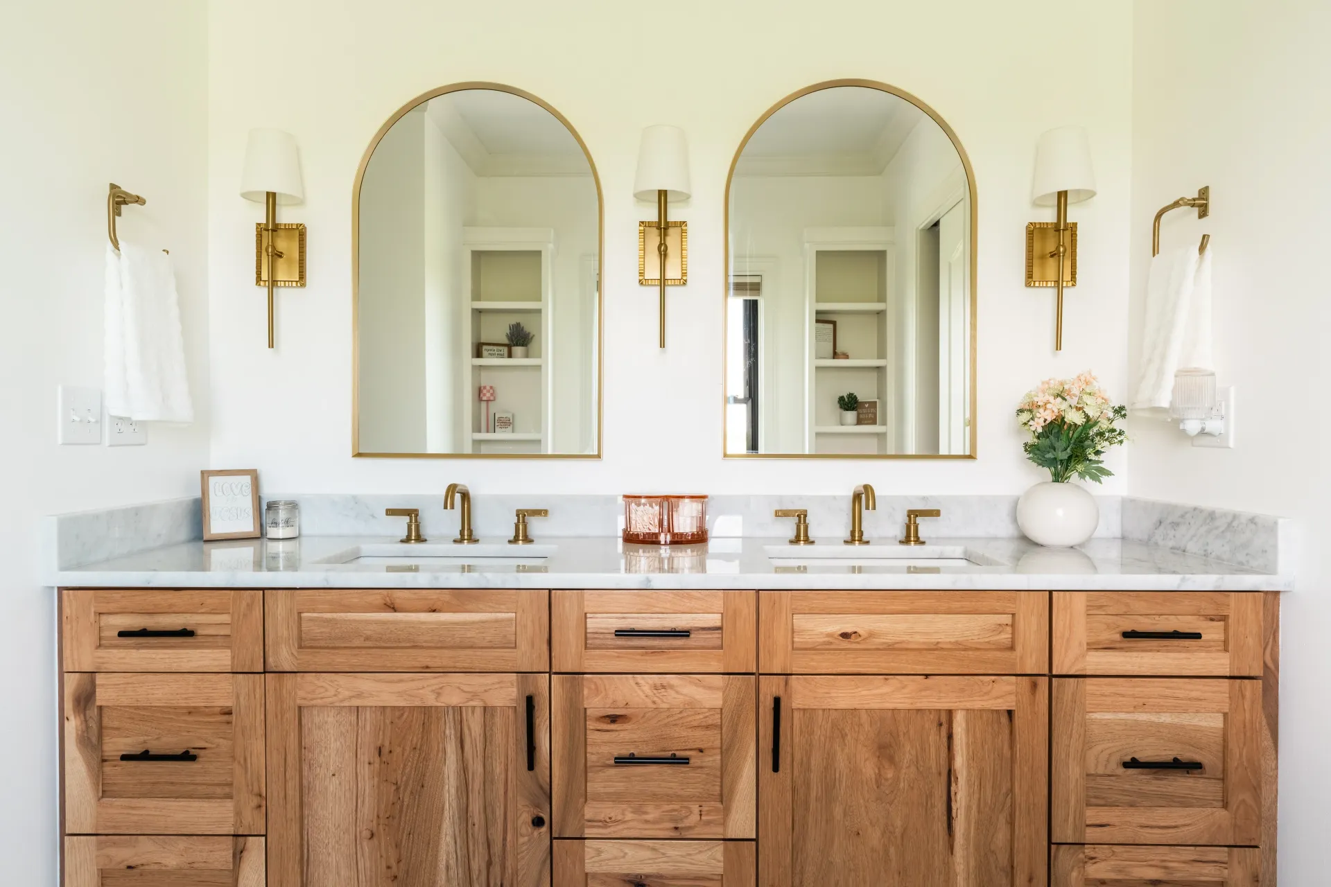 Bathroom with wood vanity, gold arched mirrors, sconces, marble countertop, and white walls.