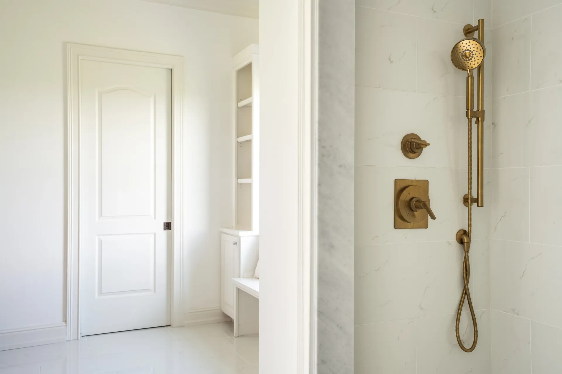 White bathroom with a gold shower fixture. A door and shelving are visible.