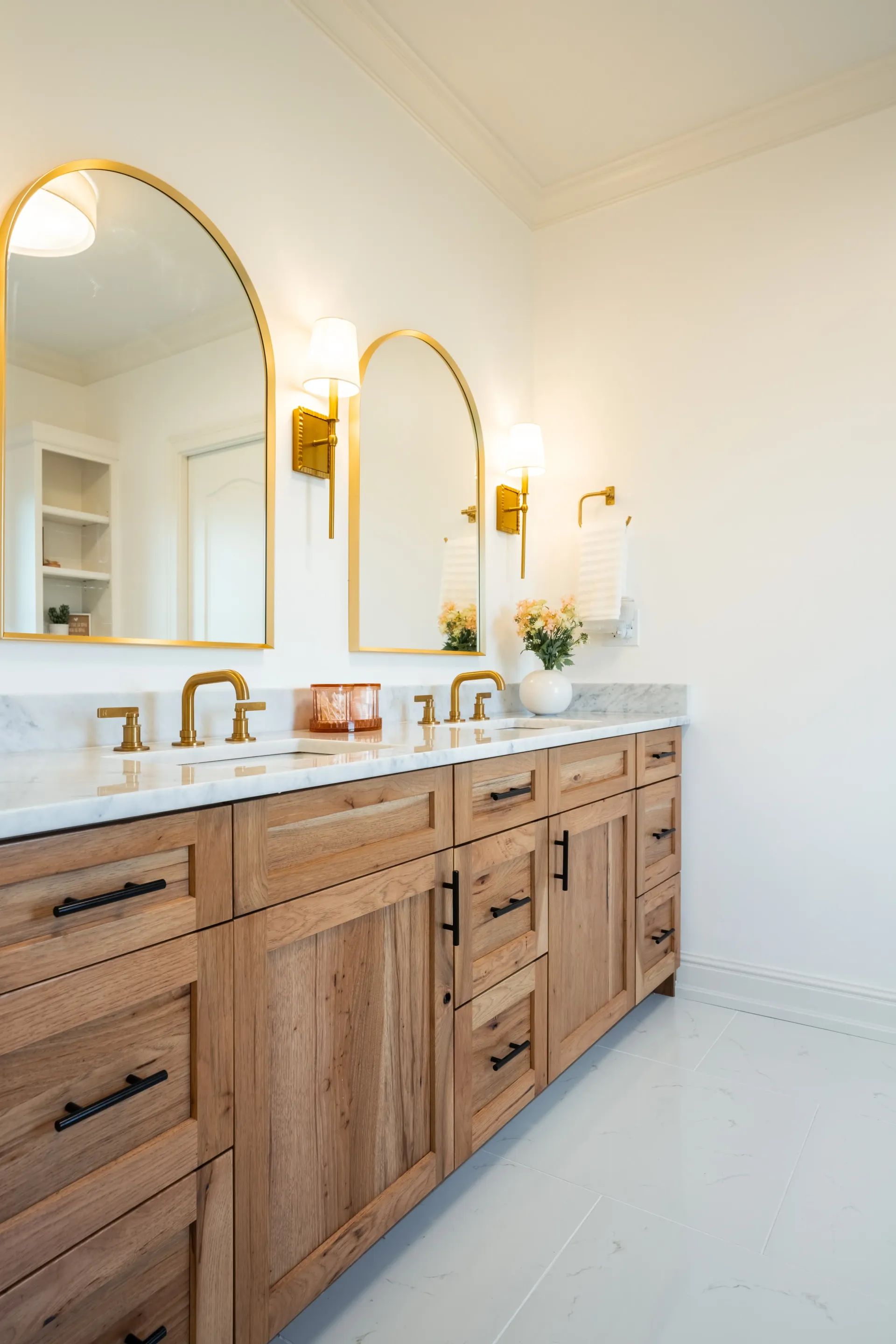 Bathroom with wooden vanity, gold mirrors, white countertops, and white tile floor.