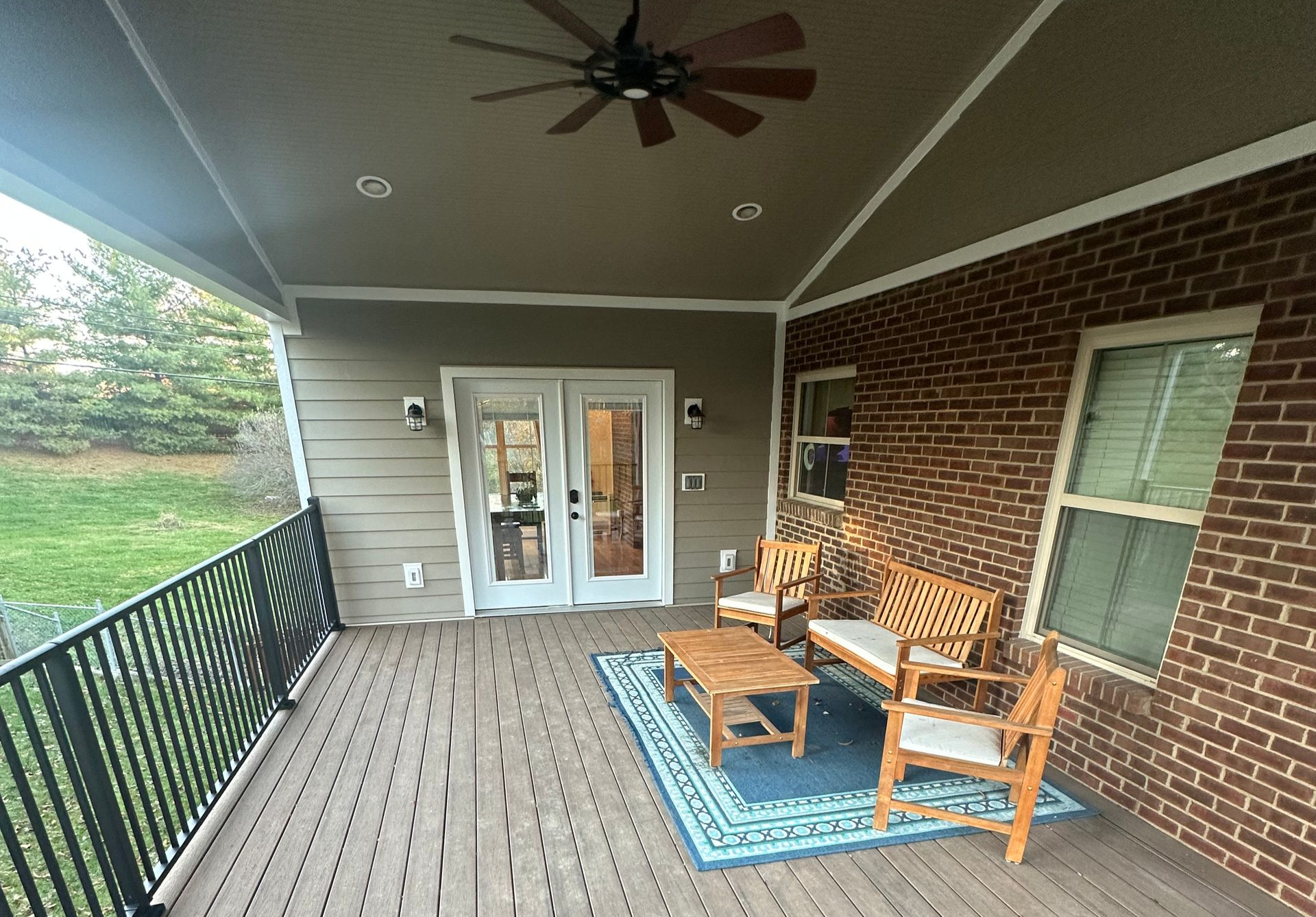 Covered outdoor deck with seating, ceiling fan, and brick wall.