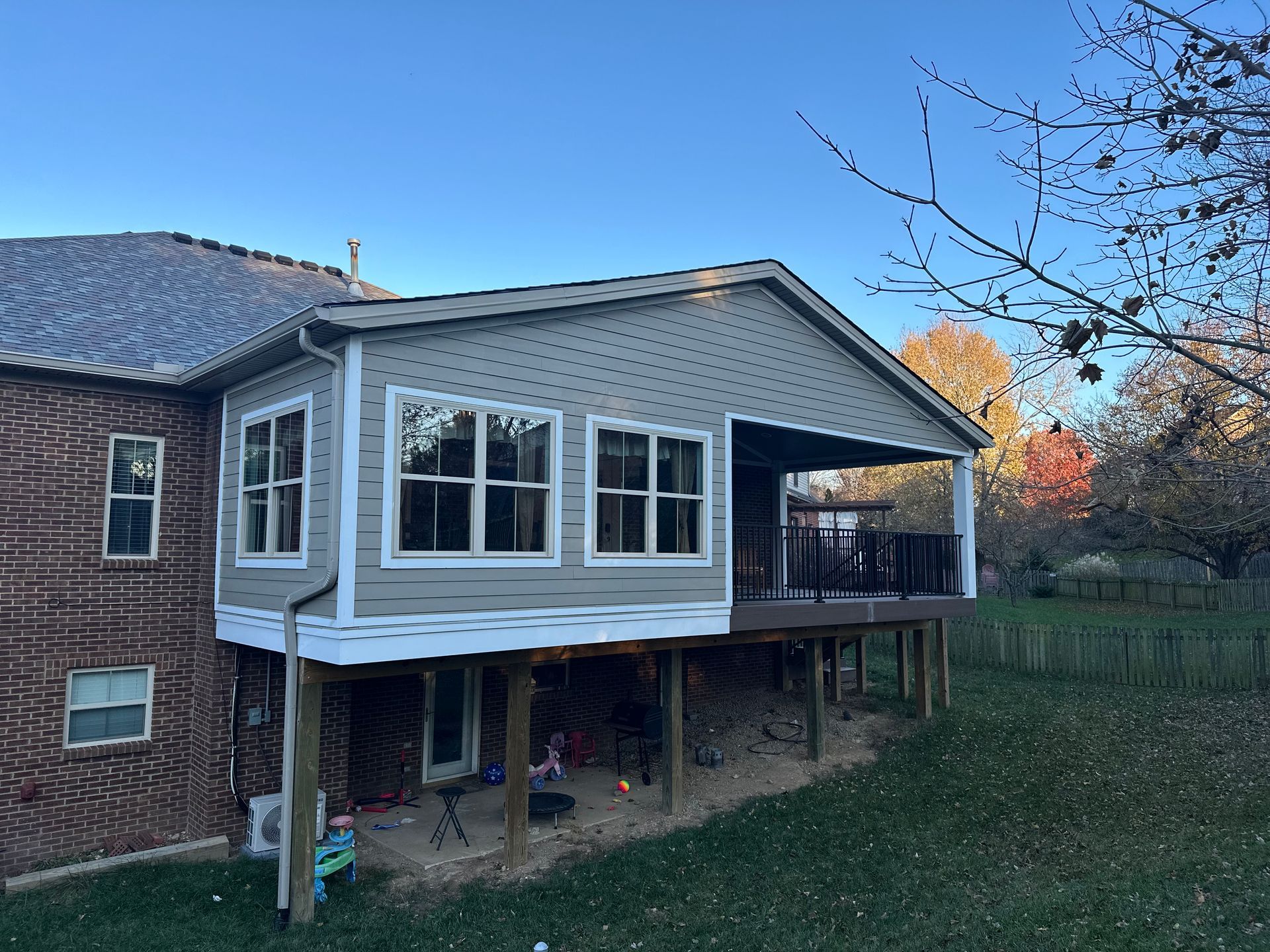 A sunroom and deck attached to a brick house. Grey siding, large windows, and a blue sky overhead.