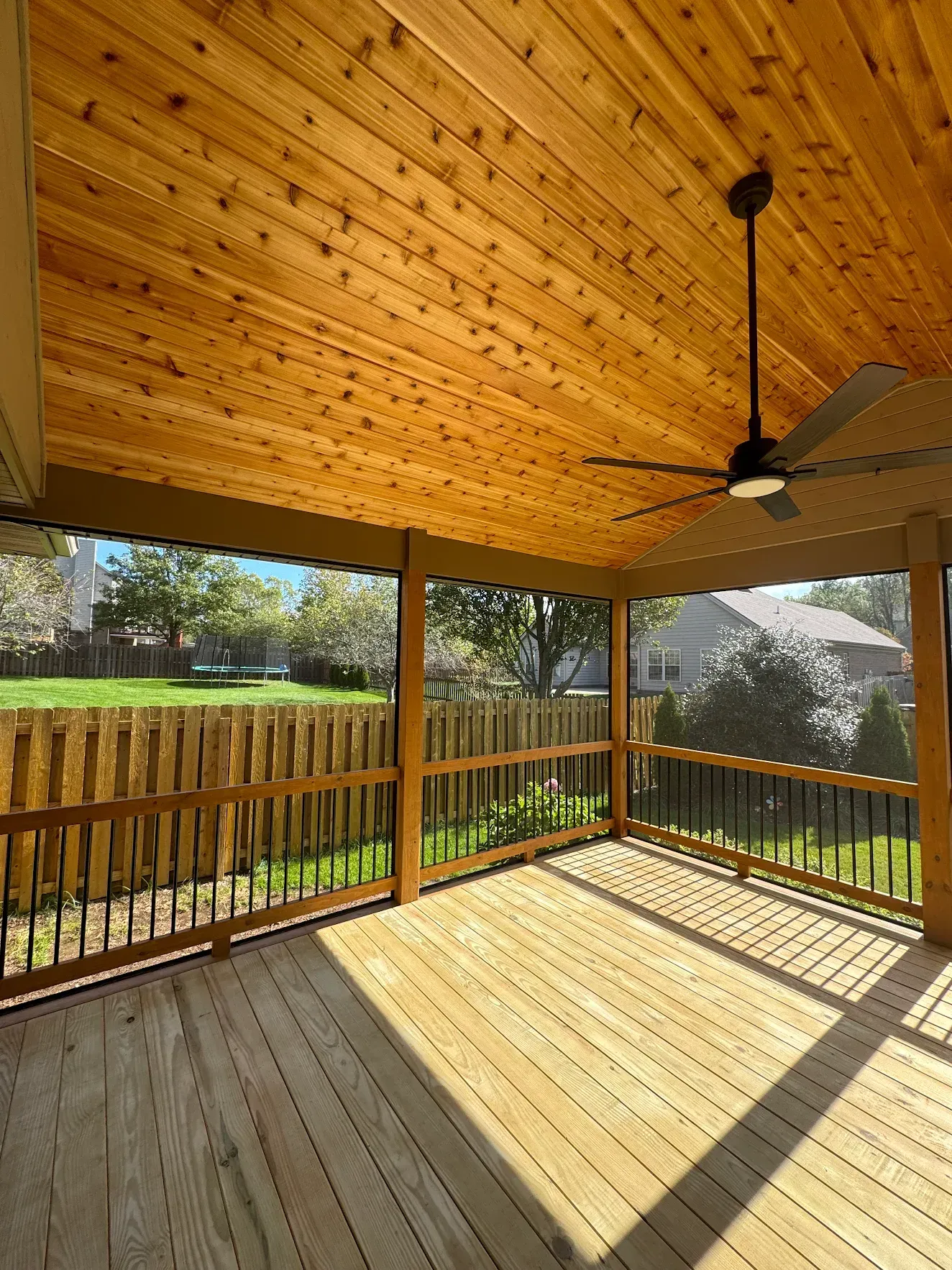 Screened-in porch with wood ceiling and deck. Black ceiling fan. View of backyard through screen.