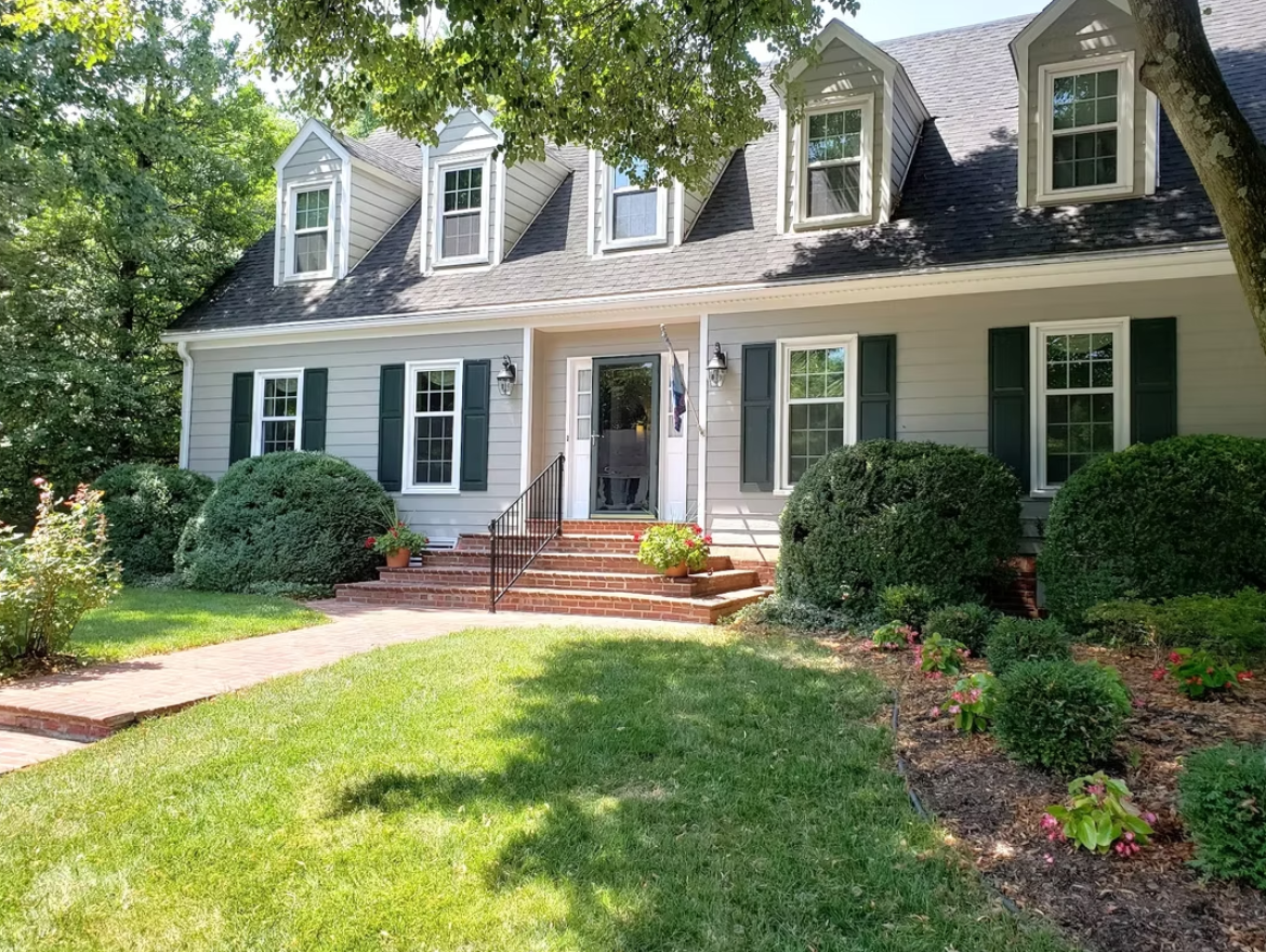 A gray house with dormers, green shutters, and a brick pathway surrounded by green bushes and grass.