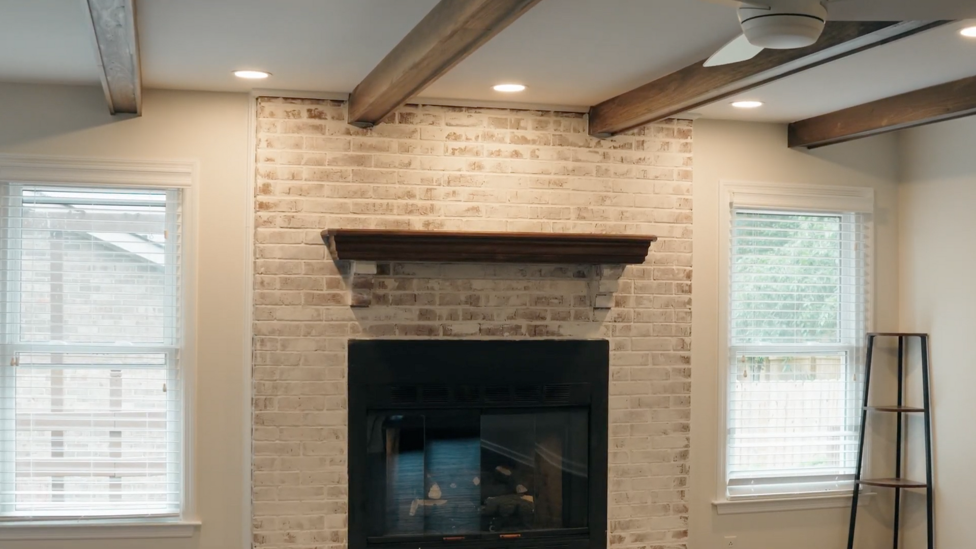 Fireplace with whitewashed brick, dark hearth, wooden mantel, recessed lights, windows, and ceiling beams.
