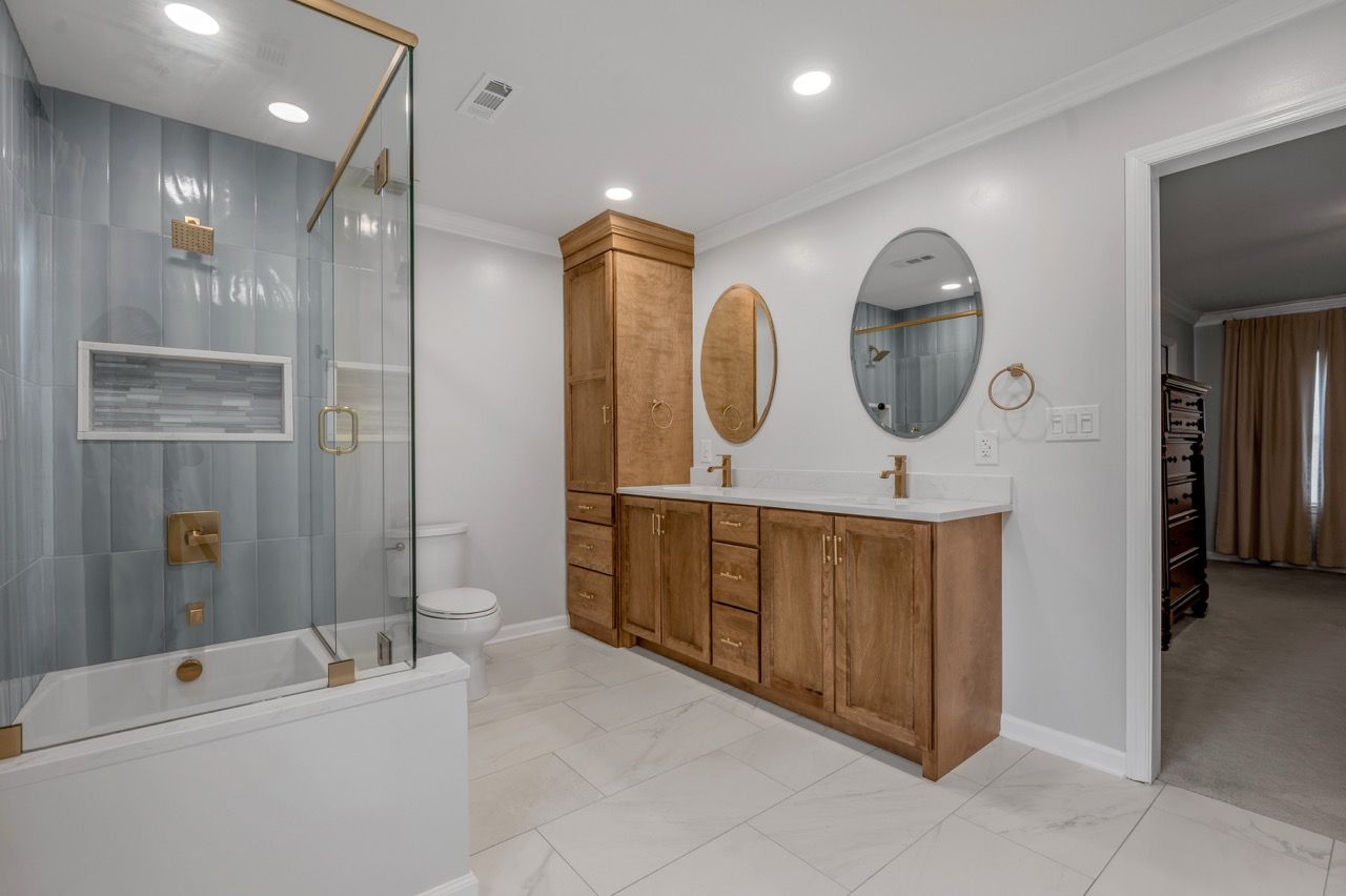 A renovated bathroom with a shower, bathtub, vanity, and a closet; featuring blue tiles, white and gold fixtures, and light-colored wood.