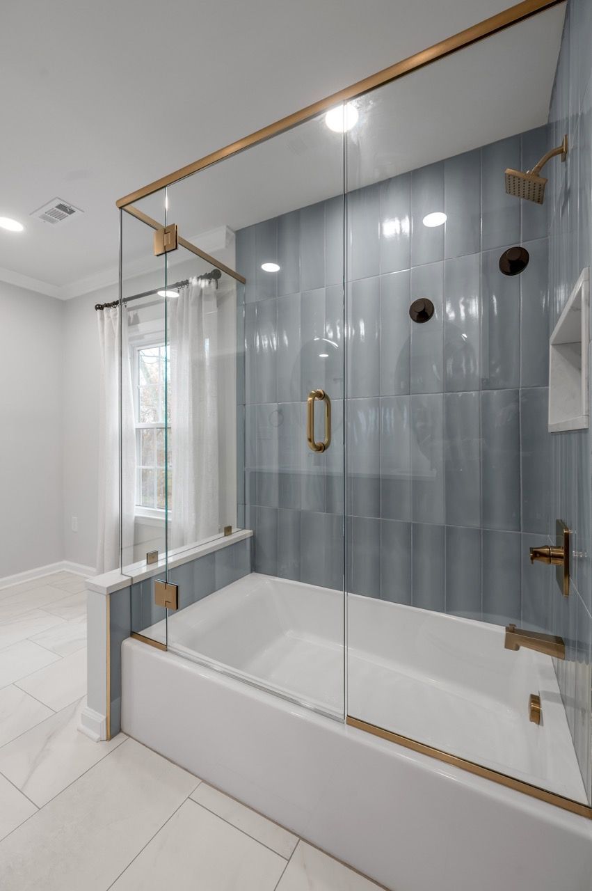 Bathroom with a white tub and blue tiled wall. Glass shower enclosure with gold fixtures.