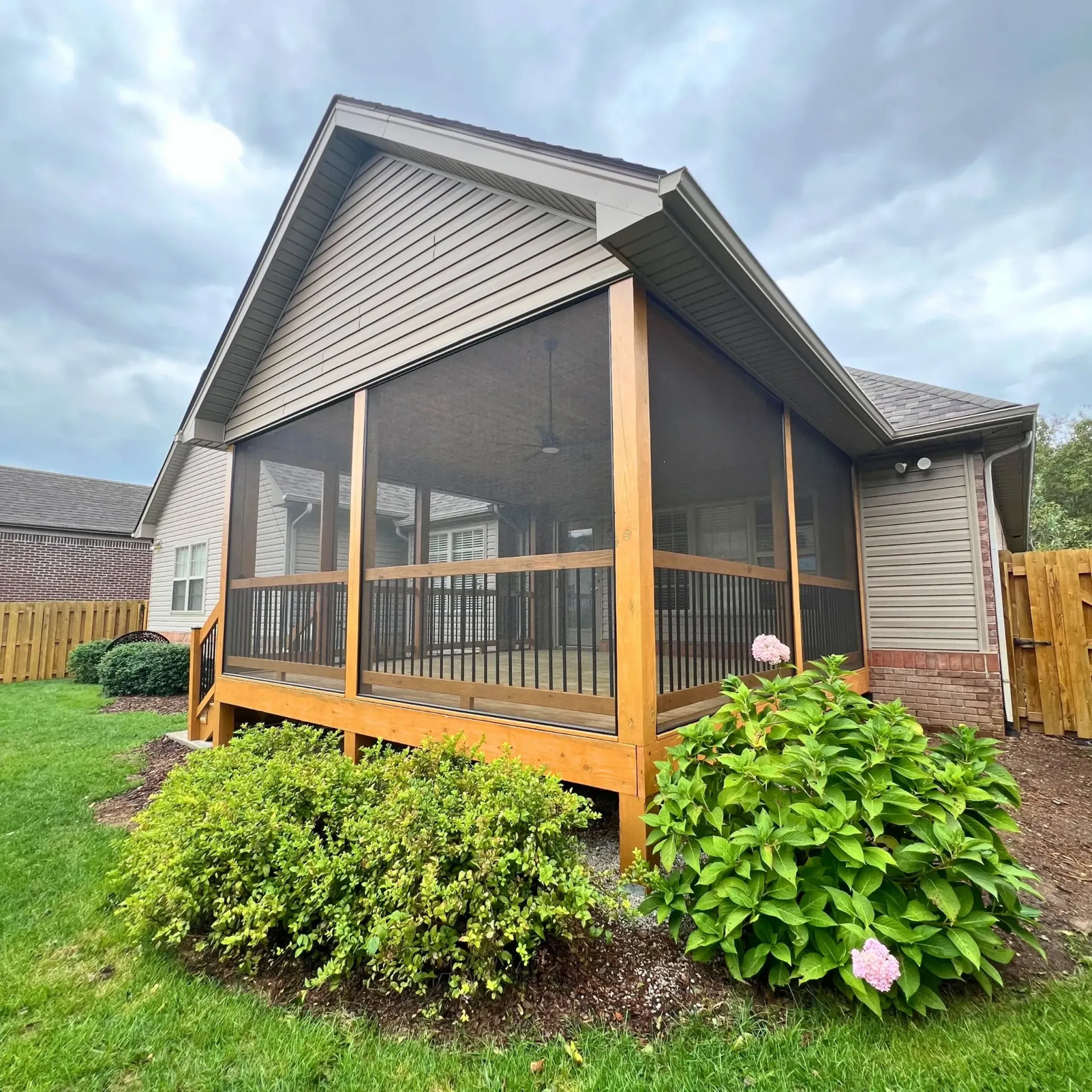 Screened porch with wooden deck attached to a house with beige siding, green bushes, and brown fence.