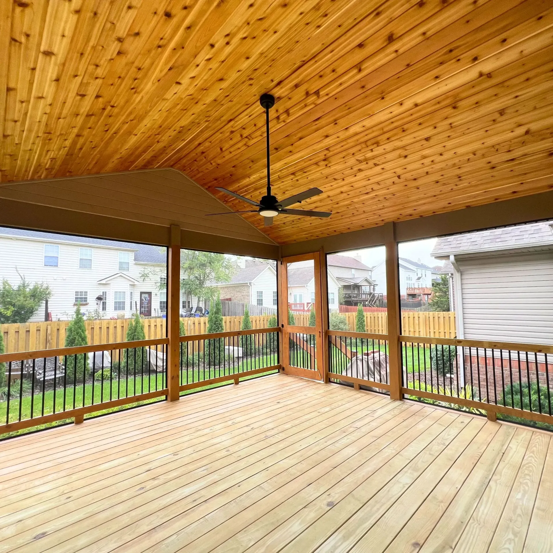 Covered wooden deck with black railings and a ceiling fan, overlooking a fenced backyard.