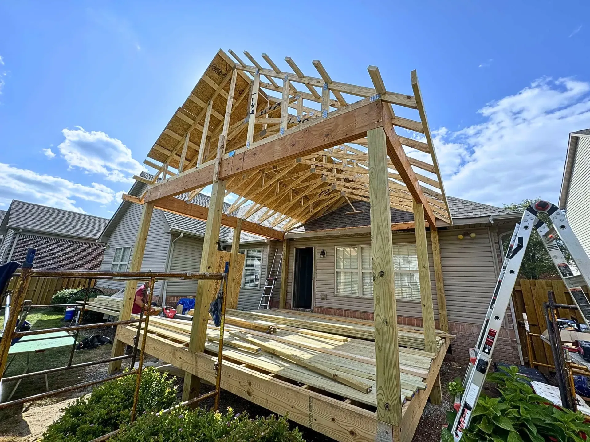 A house under construction. A wooden deck and framing for a roof extension are visible. Blue sky in the background.