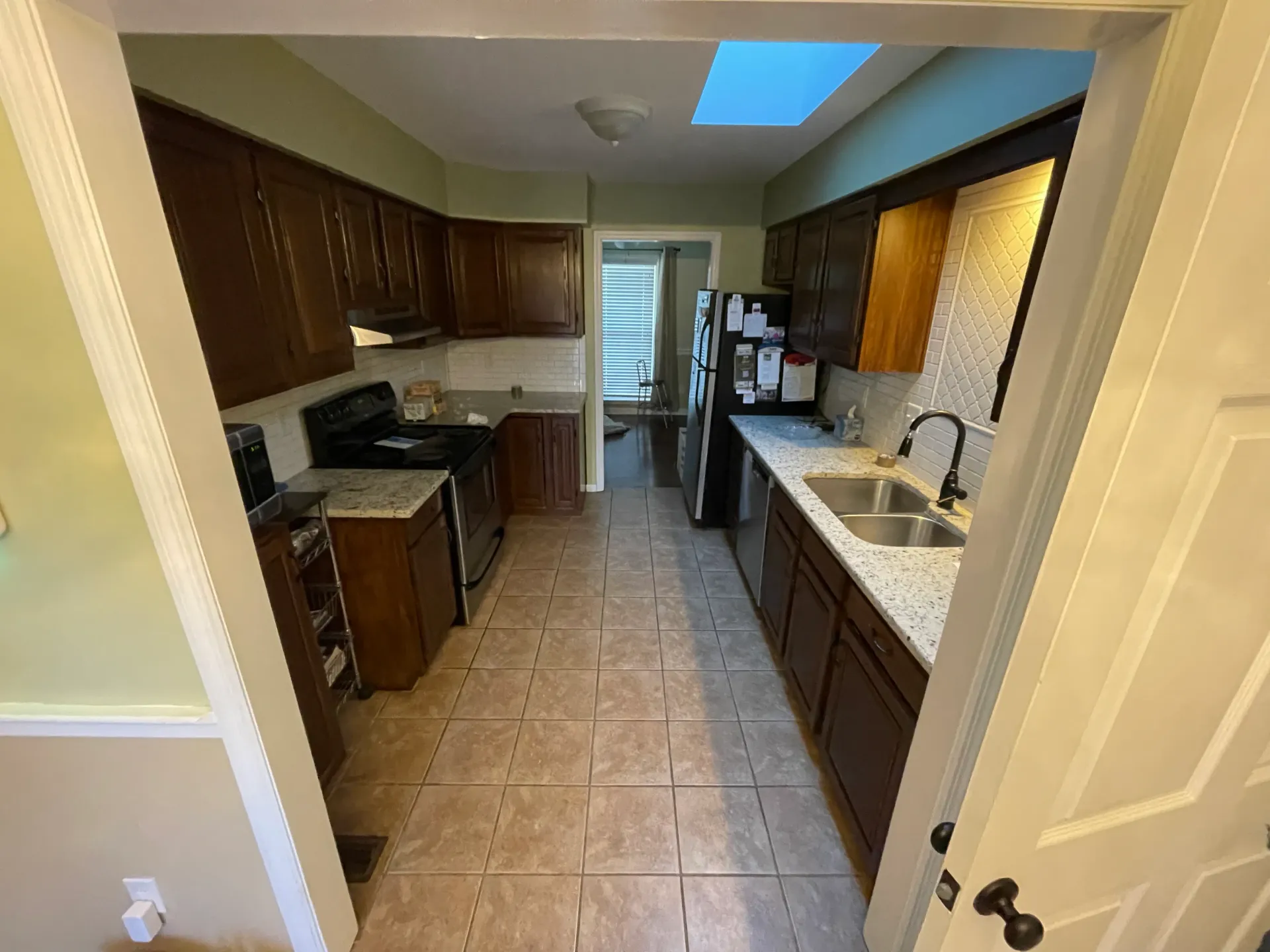 Kitchen with dark cabinets, light countertops, and tile floor, viewed through a doorway.
