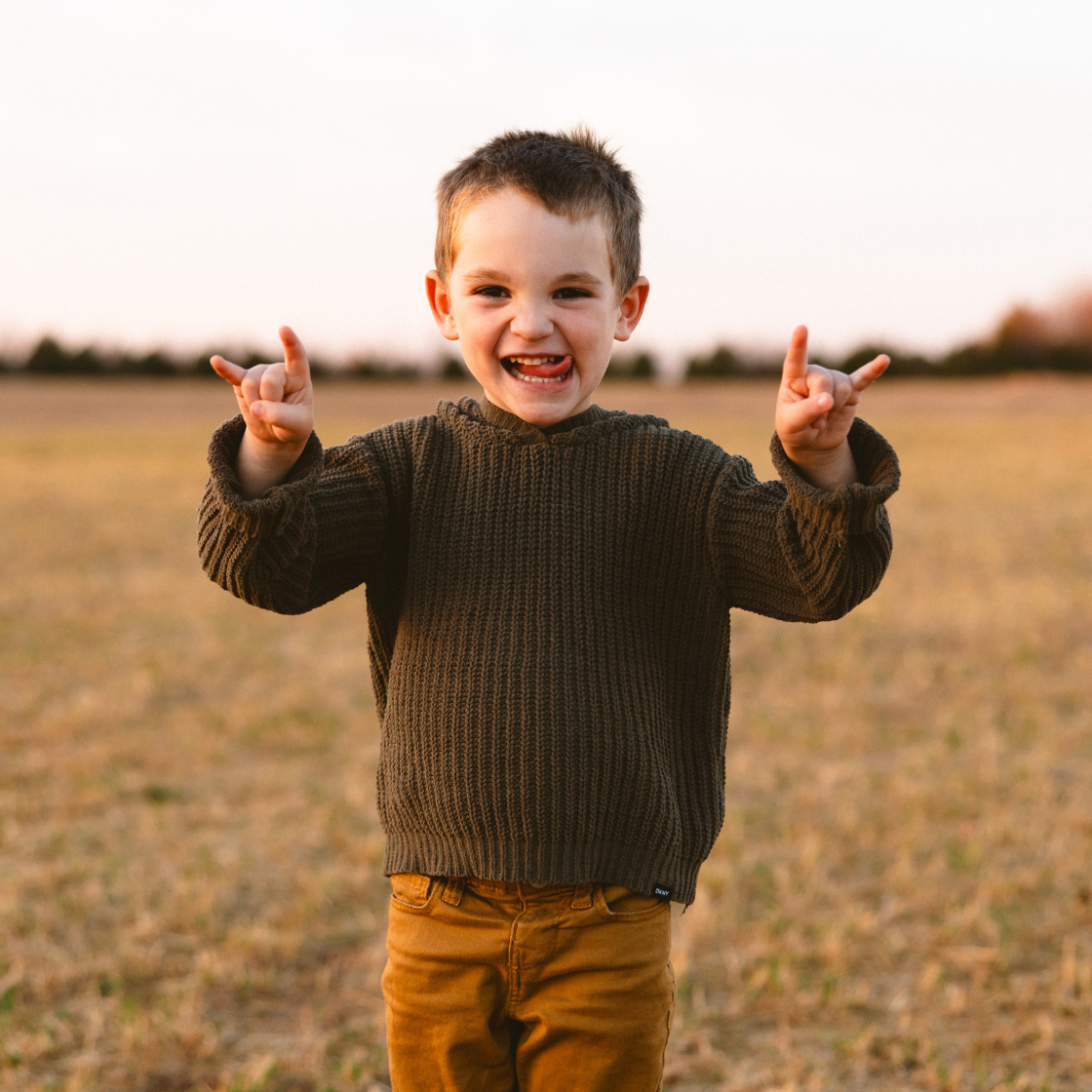 A young boy in a sweater is standing in a field making a rock and roll sign.