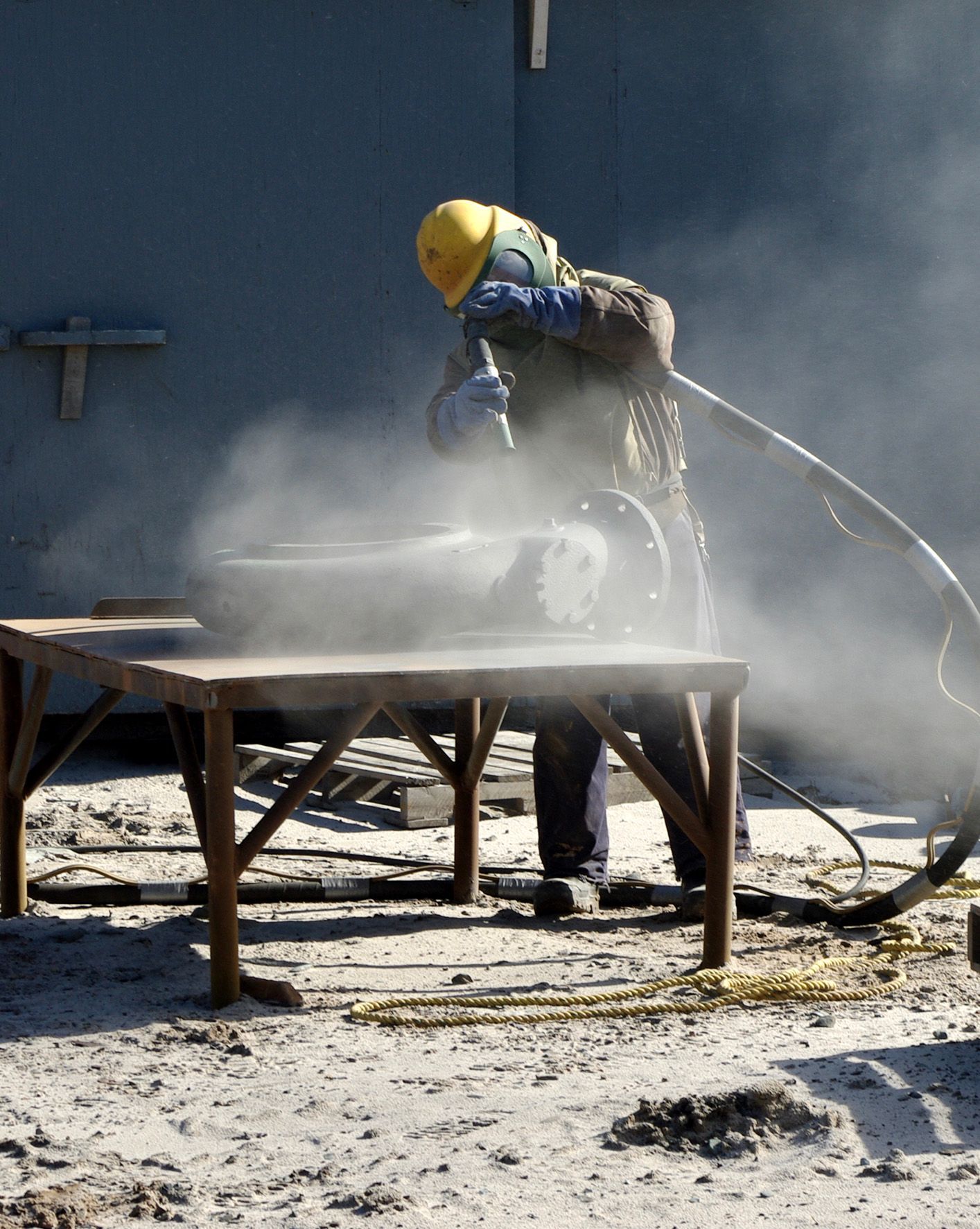 Person in protective gear sandblasting object on a wooden table, creating a cloud of dust outdoors.