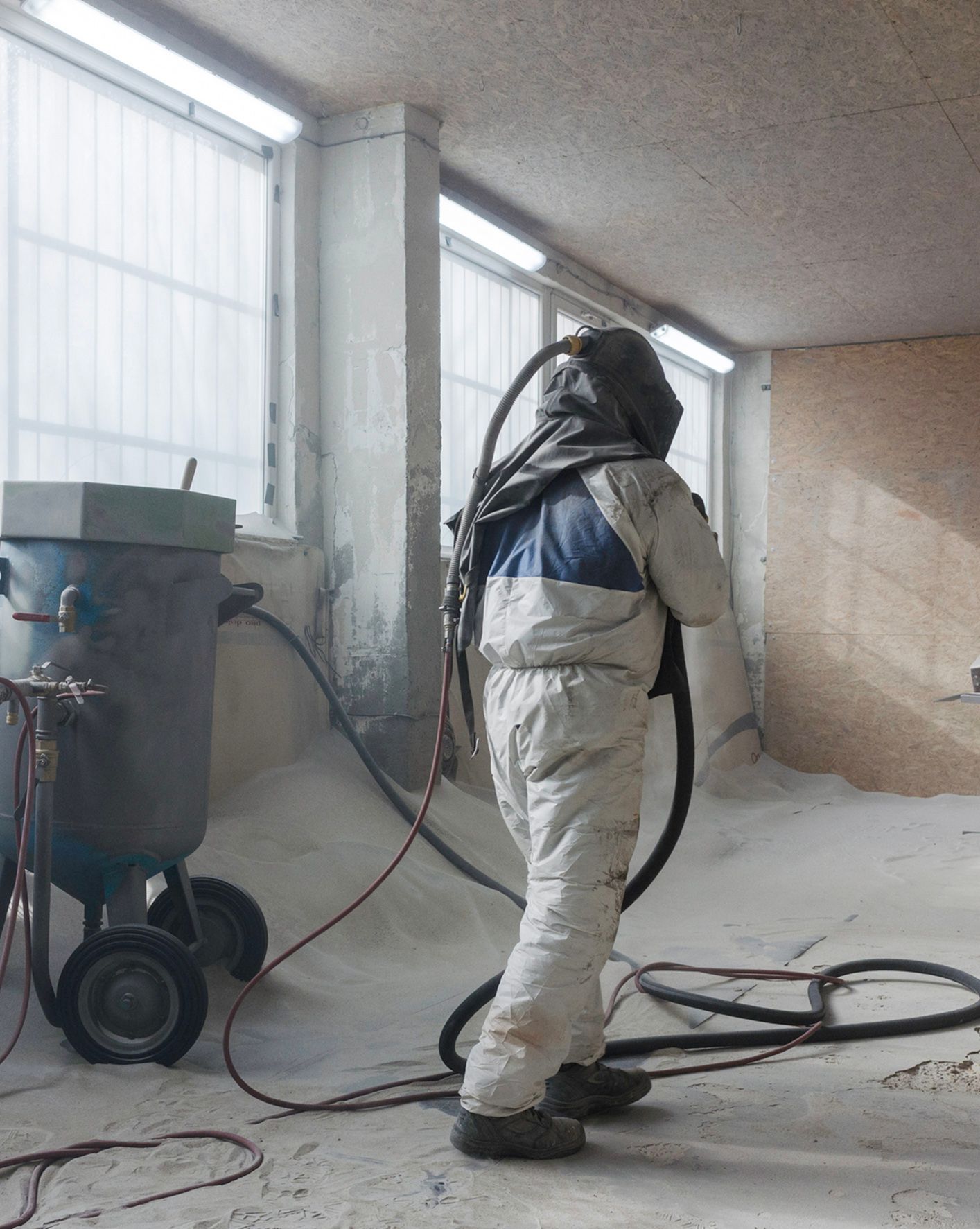 Person in protective gear sandblasting a room, connected to a tank, covered in light dust.