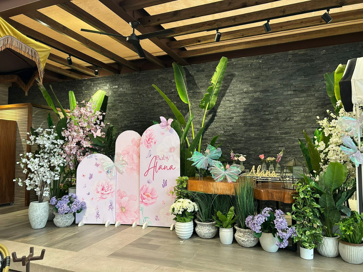 Fondo para eventos con motivos florales en tonos pastel, paneles blancos, arreglos florales y plantas en macetas bajo un dosel de madera.