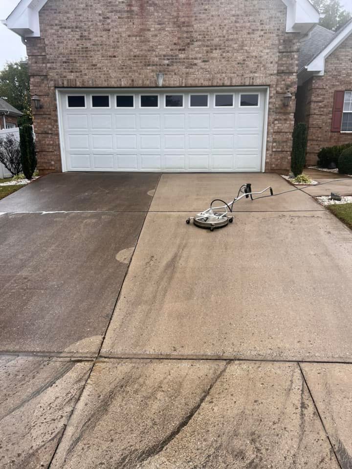Driveway being power washed; one side cleaned, the other dirty. White garage door in the background.