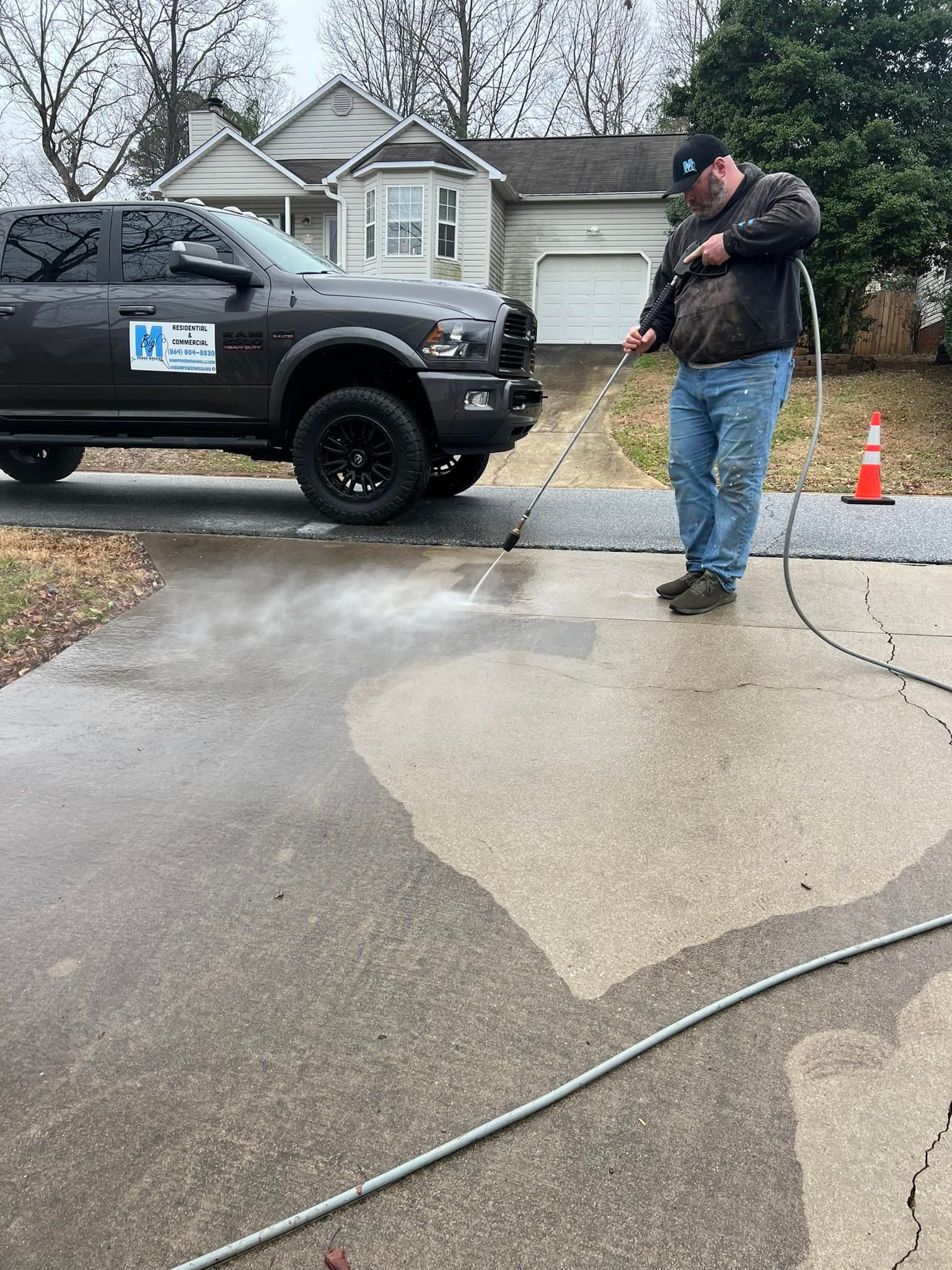 Man power washing a driveway next to a dark truck. Residential setting, overcast day.