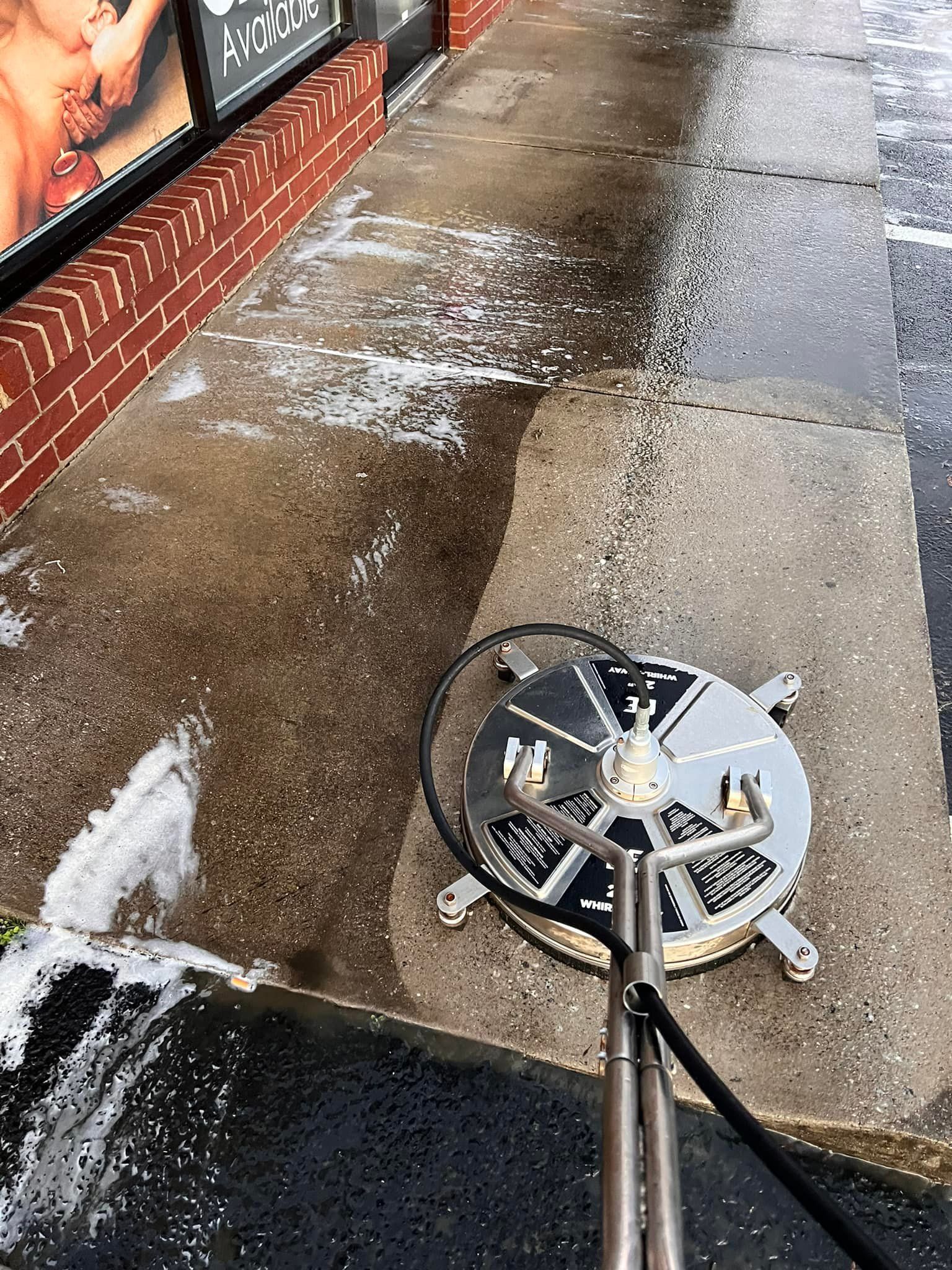 A person using a surface cleaner to wash a concrete sidewalk in front of a brick building.