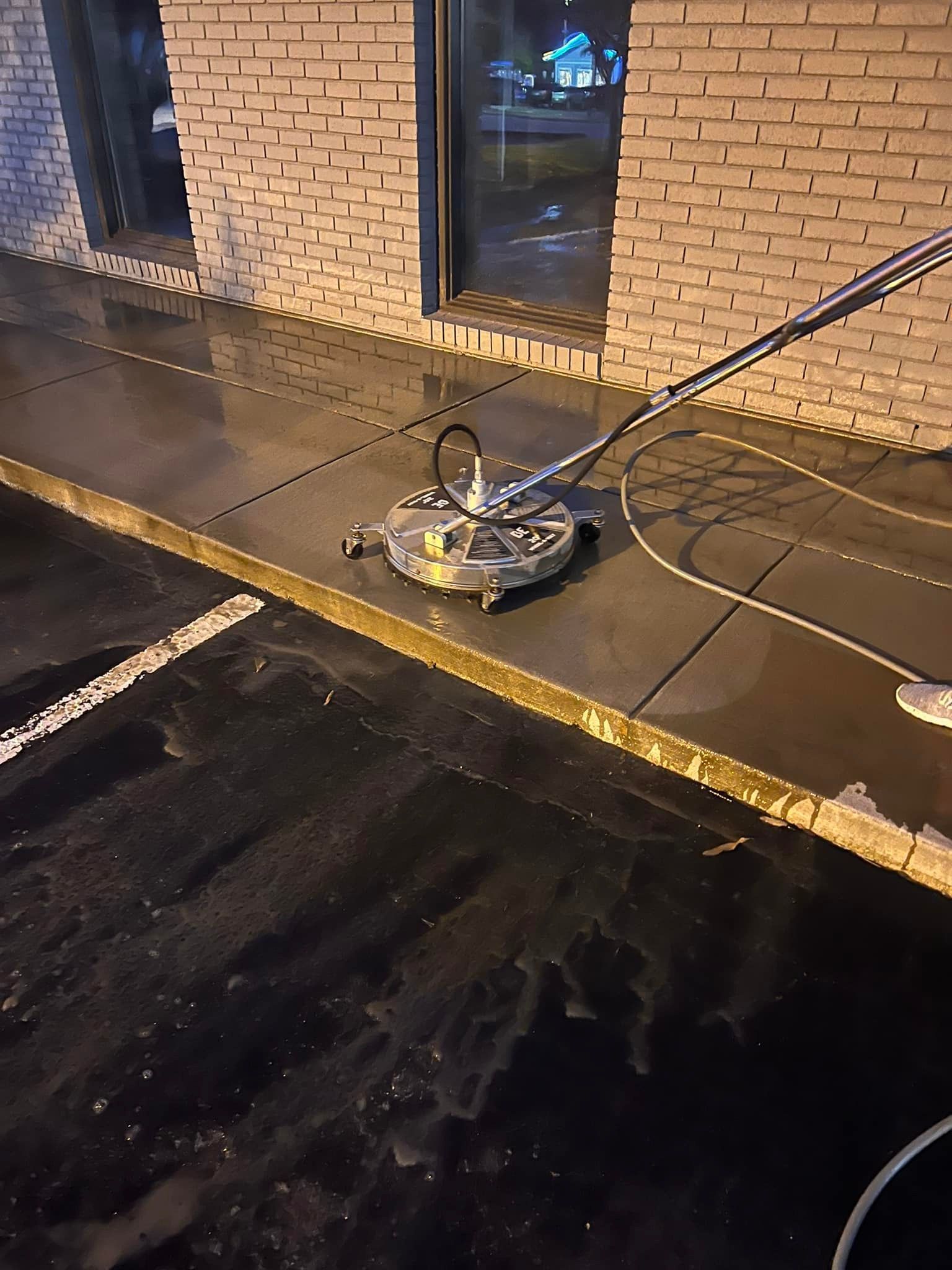 A power washer cleaning a dark, wet sidewalk next to a brick building.
