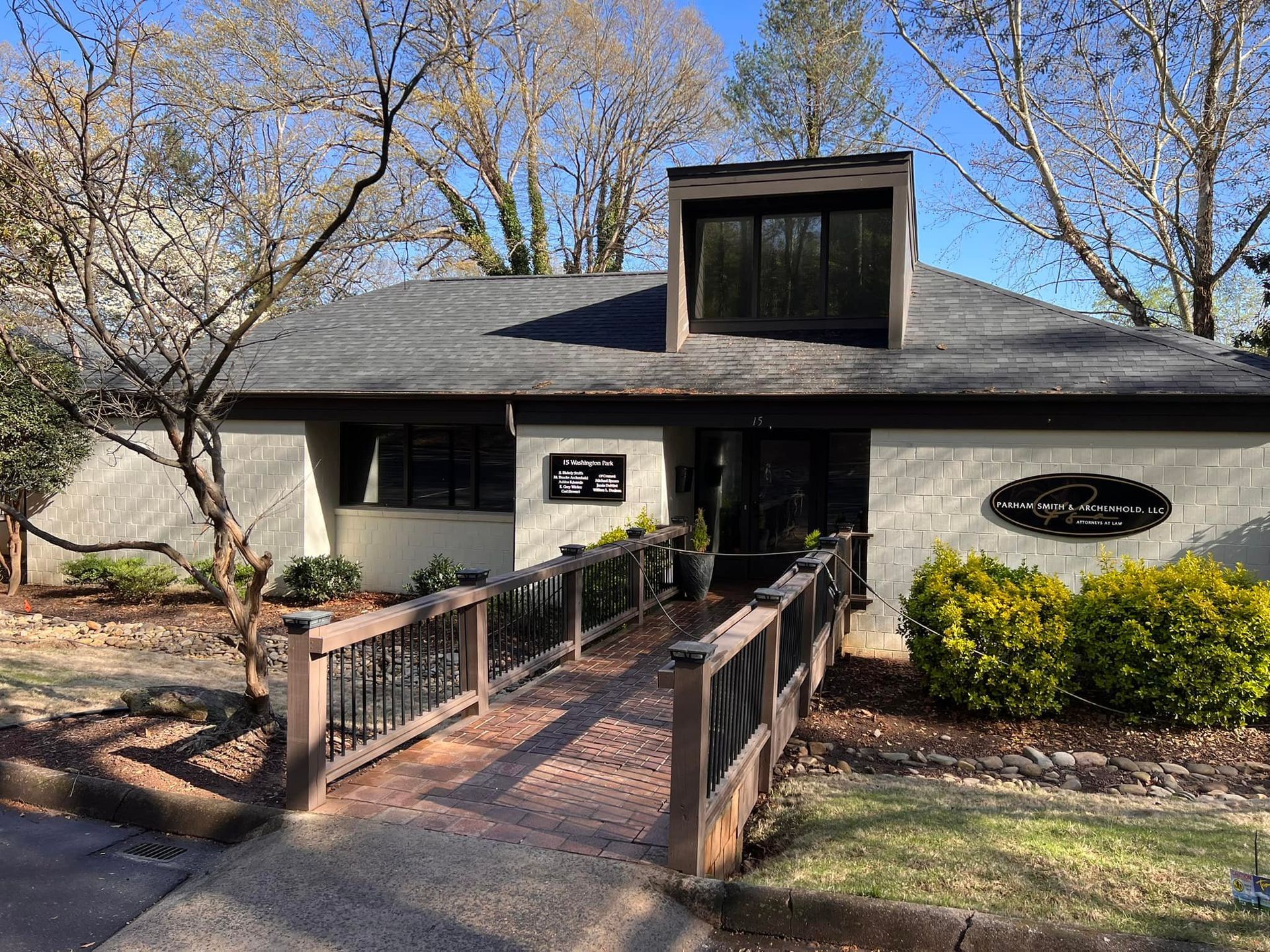 Exterior view of a one-story building with a wooden bridge leading to the entrance; trees and shrubs surround.