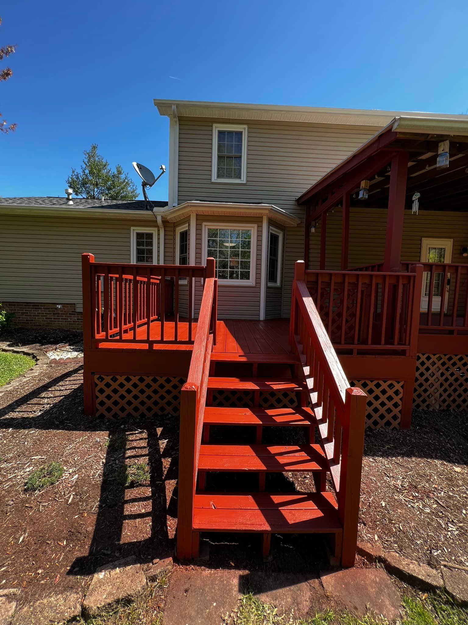 Red painted deck with stairs leading to a two-story house with tan siding and a gazebo. Blue sky overhead.