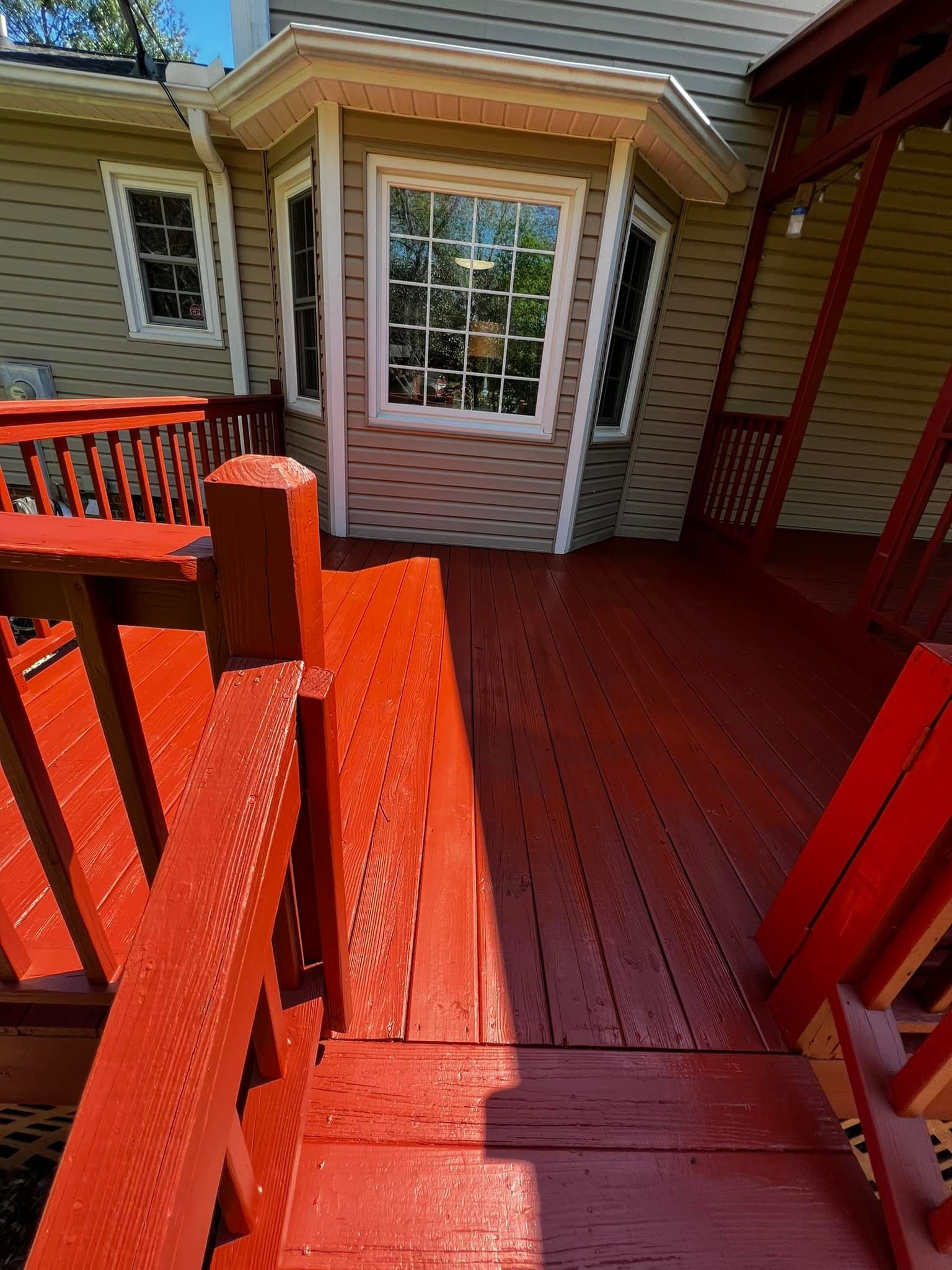 Red painted wooden deck with railing leading to a light beige house with bay windows.