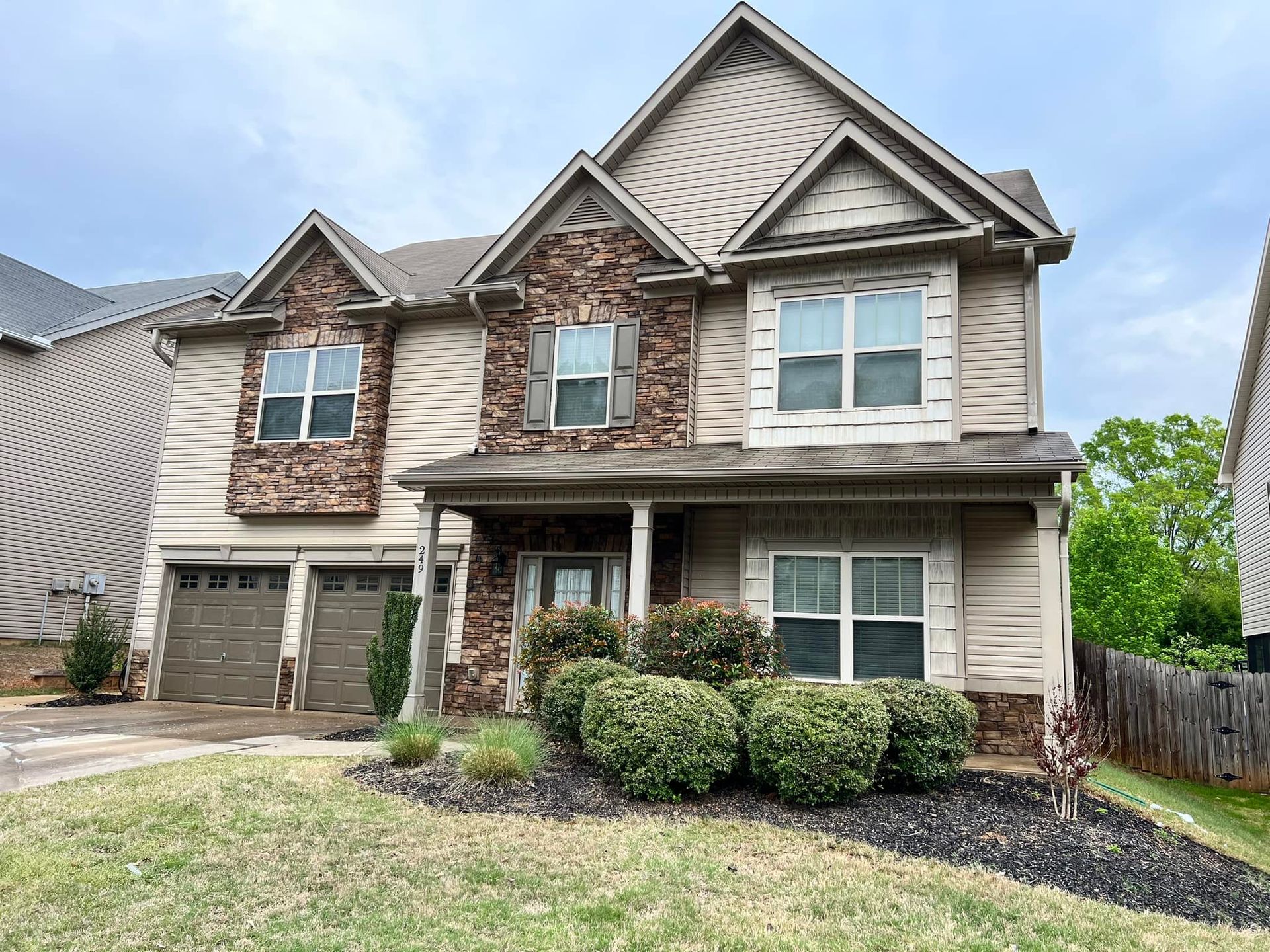 Two-story beige house with stone accents, a two-car garage, and a small front yard with bushes.