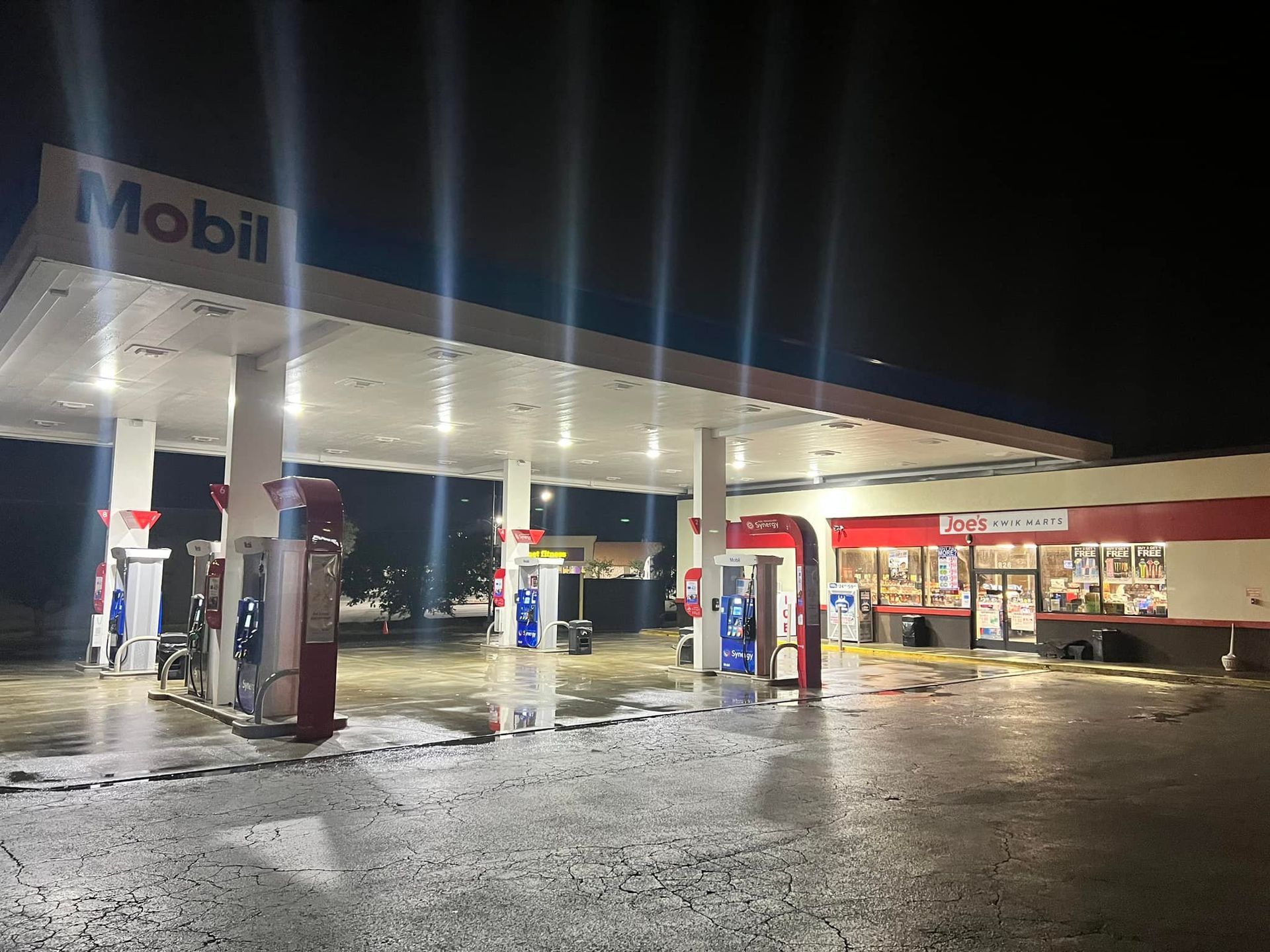 A Mobil gas station at night, with illuminated pumps and convenience store; wet pavement reflects lights.