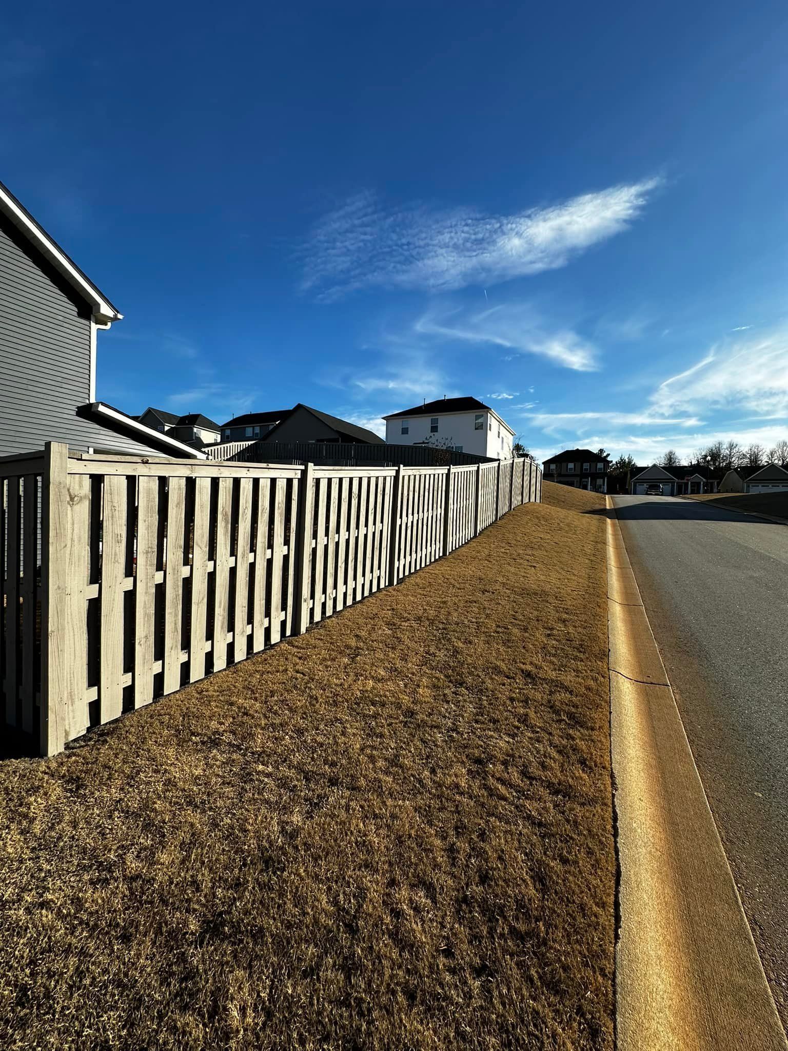 Wooden fence along a road, houses in background, blue sky.