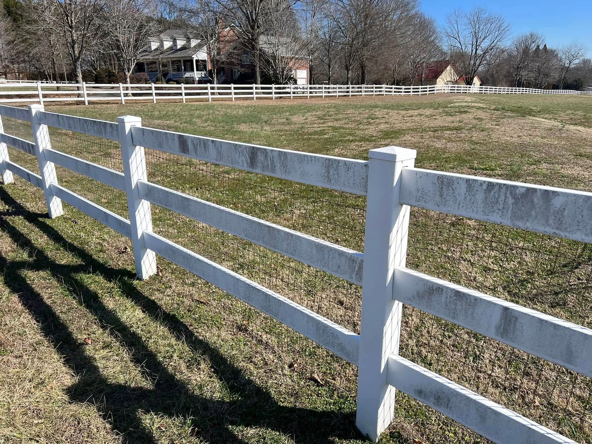 White, weathered three-rail fence in a grassy field, with a house in the background on a sunny day.