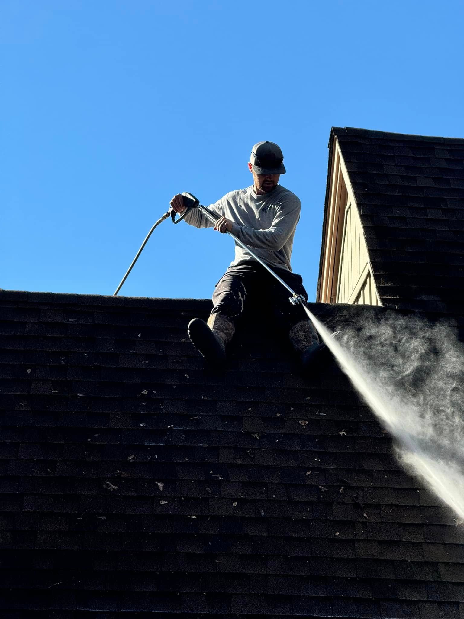 Person pressure washing a dark shingle roof with a blue sky background.