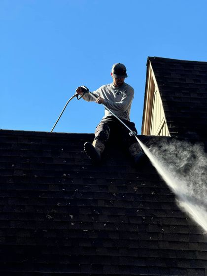 Person power washing a dark shingled roof under a clear blue sky.