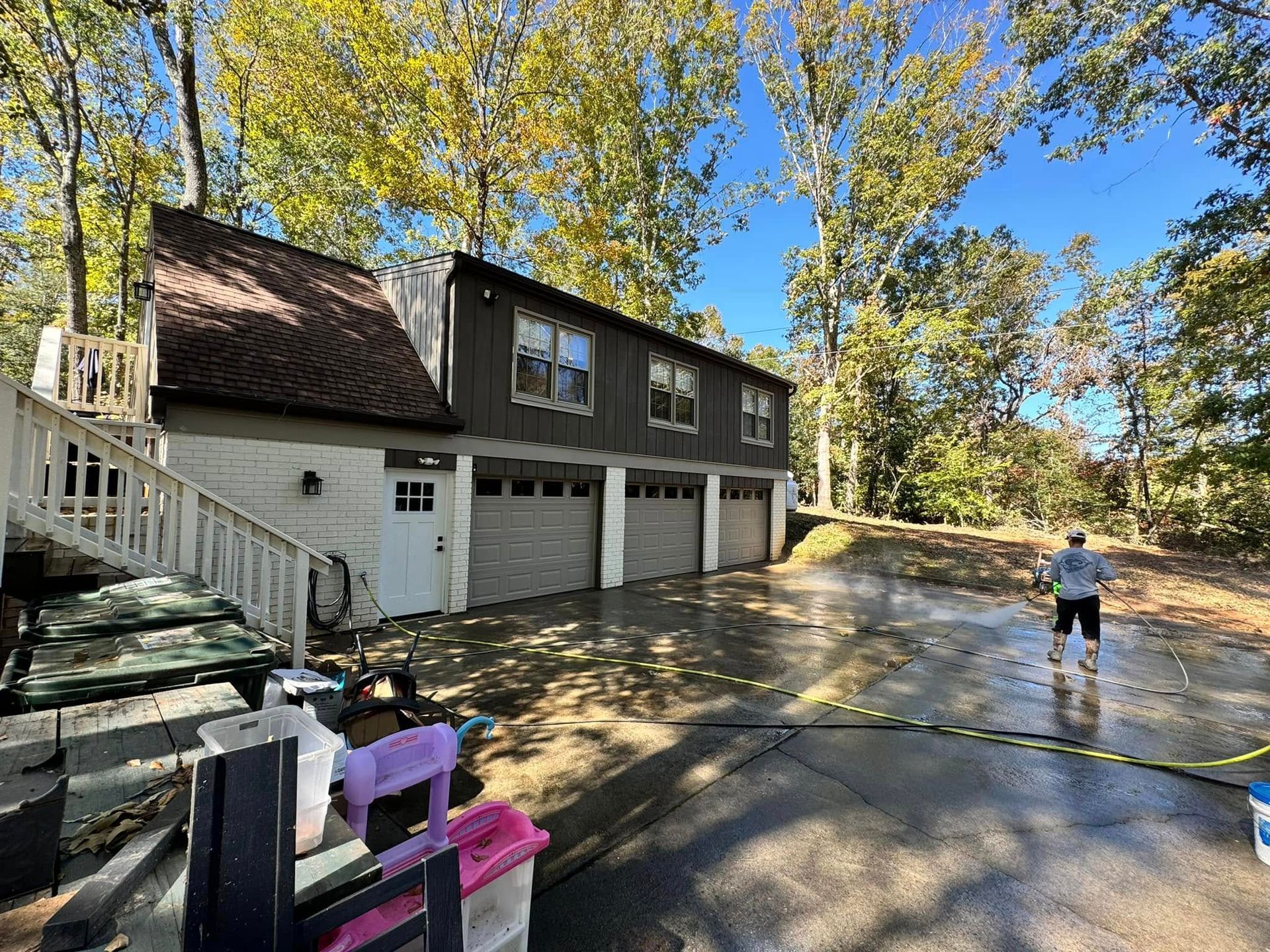 A person power washes a driveway in front of a three-car garage and a house with a deck; autumn trees surround.