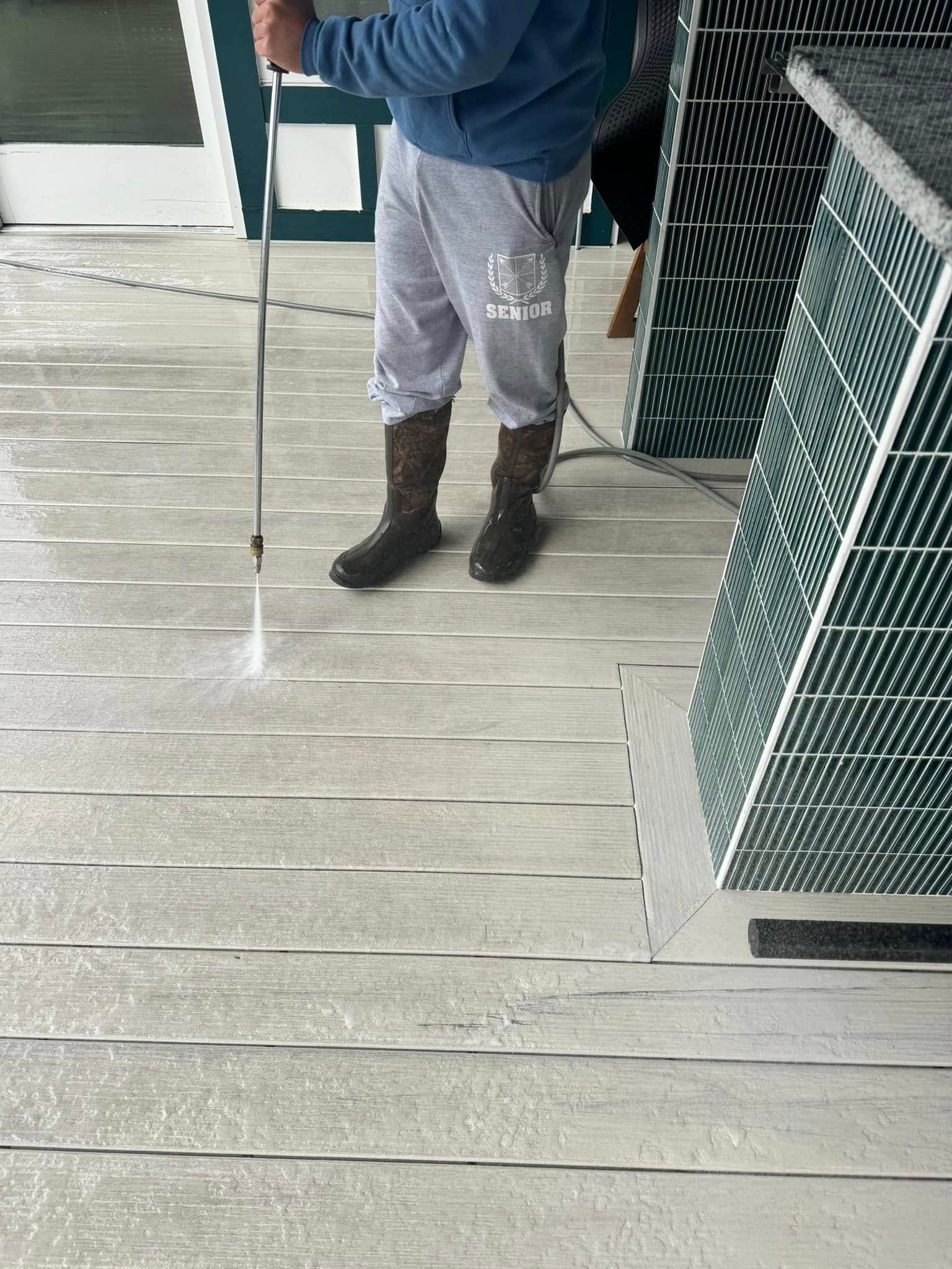 Person power washing a gray wooden deck, wearing boots and sweatpants.