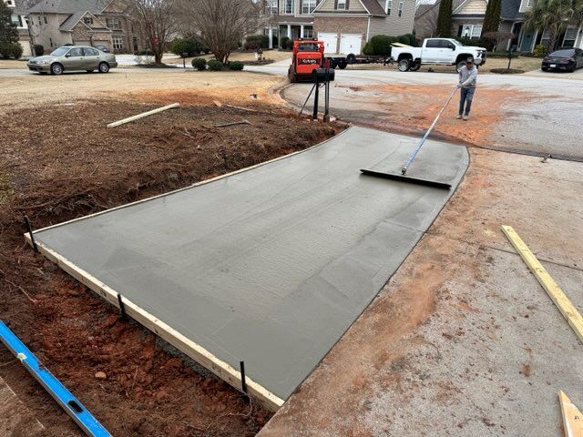 Freshly poured concrete driveway being smoothed; person raking, setting is residential.