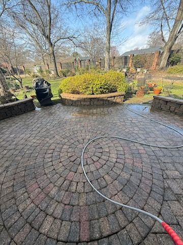 Circular brick patio being cleaned with a hose in a backyard with landscaping and trees.