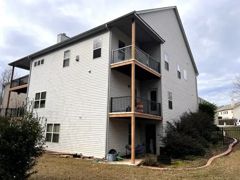 Two-story house with multiple balconies and gray siding, brown support posts. Cloudy sky, backyard setting.